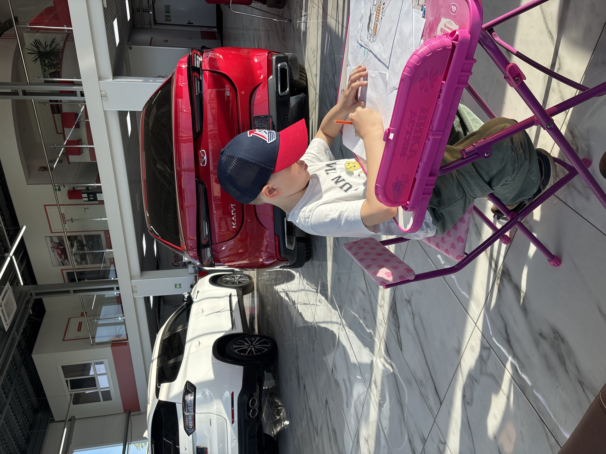 The image captures a candid moment in what appears to be a brightly lit car dealership showroom in Yaroslavl, Russia. The main subject is a young boy, possibly around 5-7 years old, engrossed in drawing or coloring. He wears a white t-shirt with partially visible text, green pants, and a dark blue and red baseball cap with an embroidered logo. He is seated on a small, vibrant pink and purple children's chair with an integrated desk, using an orange pencil or crayon on a piece of paper.
The setting is a modern, spacious indoor environment with a polished white tiled floor reflecting sunlight and structural elements. Several vehicles are on display. Directly behind the child is a striking red SUV, identified by "KAIYI" on its rear badging and a numerical plate "1600-00". Further into the showroom, a white SUV, with "CHERY" visible on its rear, is also prominently displayed. These are consistent with vehicles sold in Russia.
Notable details include the strong sunlight casting distinct shadows on the floor, indicating daytime. The children's desk, despite being used by a boy, bears Cyrillic text "НИКА АКТИВ" (Nika Activ) and "Маленькая принцесса" (Little Princess), along with a "NIKA KIDS" logo. In the background, modern architectural features like white pillars, glass partitions, and red accent walls create a clean aesthetic. A red fire extinguisher and an exit sign are also visible, adding to the typical car showroom environment. The child appears focused and calm, enjoying his creative activity amidst the commercial setting.