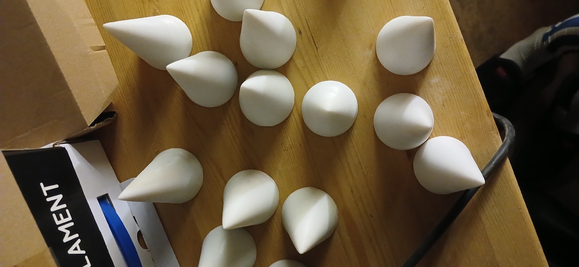 The image shows a collection of white, cone-shaped objects arranged on a light-brown wooden surface. Adjacent to the objects on the left side of the image is a cardboard box with "FILAMENT" printed on its side. The surrounding environment appears to be indoors, with a glimpse of a dark-colored garment or object visible in the background to the right. There is no verifiable information to determine that the photograph was taken in Moscow, Russia.