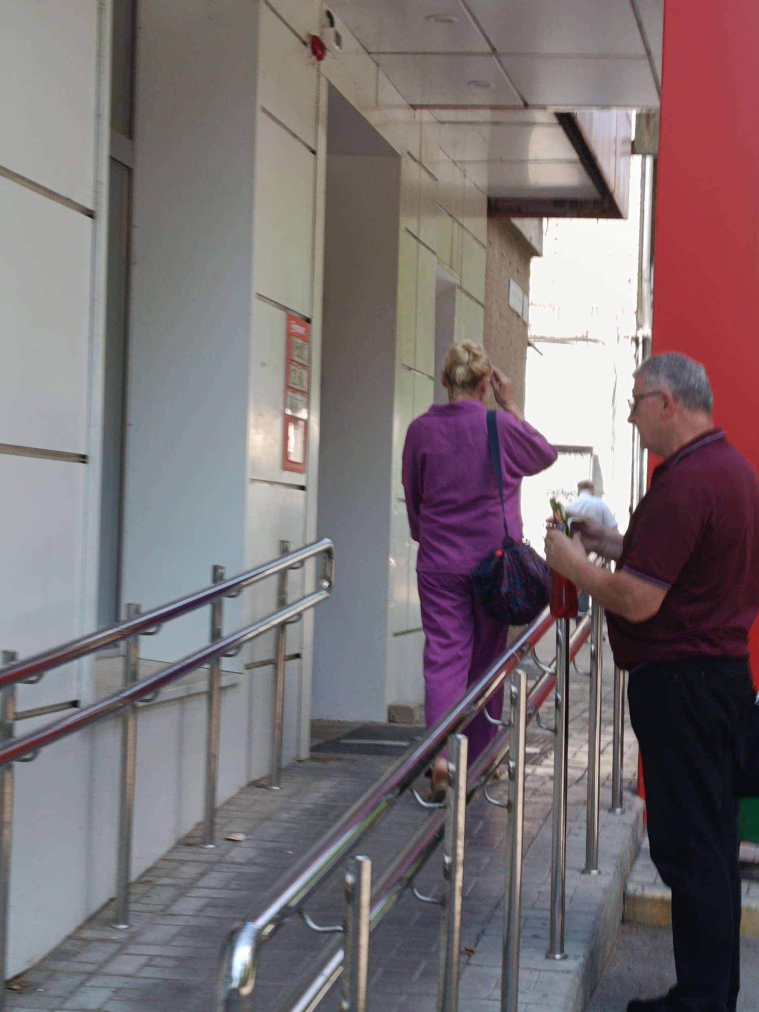 This is a medium-shot, exterior photograph taken during daylight hours. In the foreground, a man with graying hair and glasses, wearing a maroon polo shirt and black pants, stands to the right. He holds a bottle with a red liquid and a green object, possibly a straw, in his right hand, seemingly interacting with the bottle. A ramp with stainless steel railings leads up to a building entrance. Behind the man and on the ramp, a woman wearing a bright purple pantsuit with her hair in a bun walks away from the camera. She has a dark blue and patterned bag slung over her left shoulder. The building facade is white and light gray, with some red accents. The ground is paved with light-colored bricks. The environment appears to be a commercial area, possibly a shopping center or a similar establishment. Based on visual cues such as building architecture and the presence of a ramp, this scene could be located in Saratov, Russia.