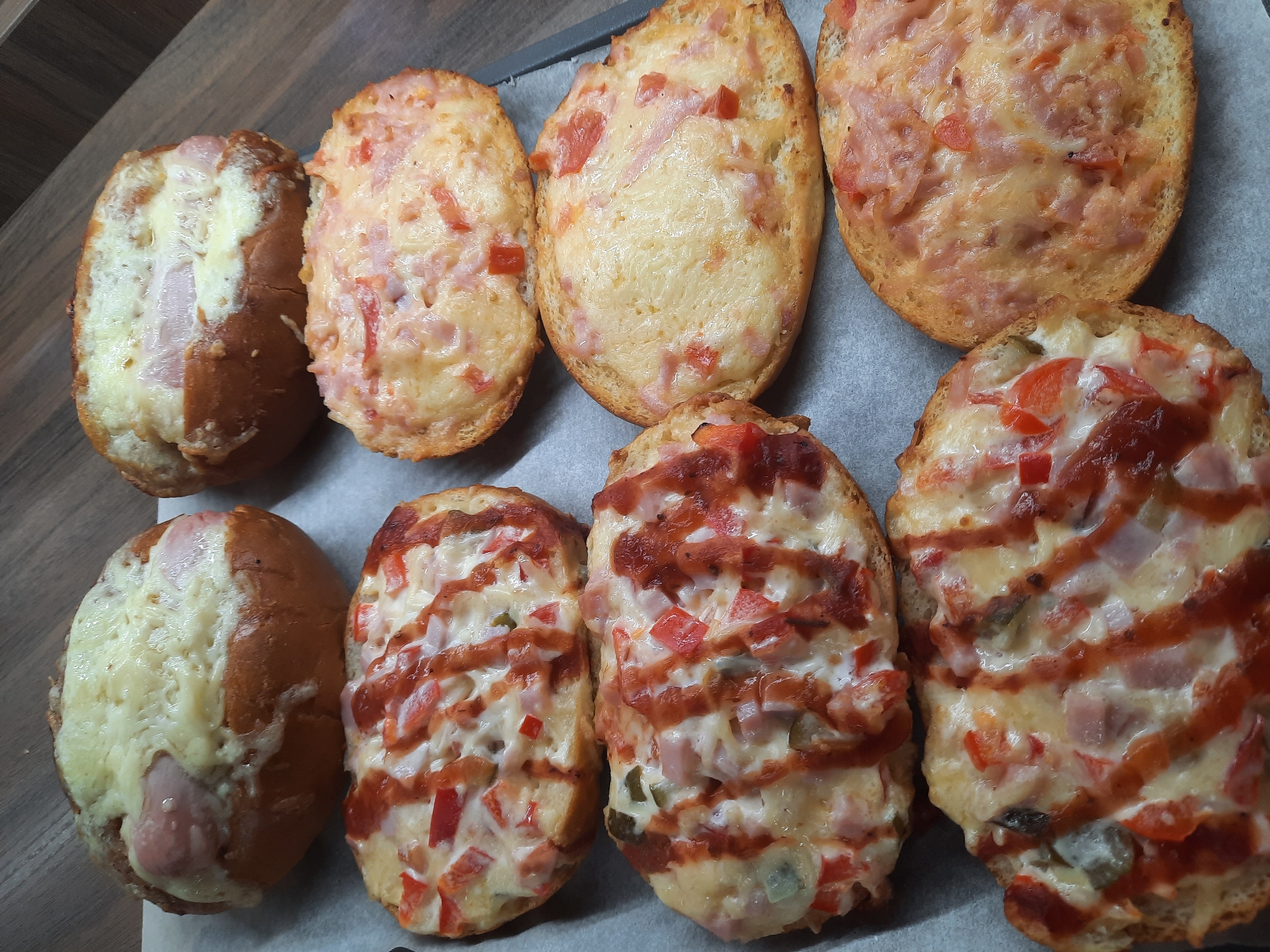On a parchment-lined baking sheet, nine baked open-faced sandwiches are arranged. The sandwiches appear to be topped with cheese, ham, and possibly other vegetables like diced red peppers and pickles. Some have a drizzle of sauce, possibly ketchup. The baking sheet sits on a wooden surface.