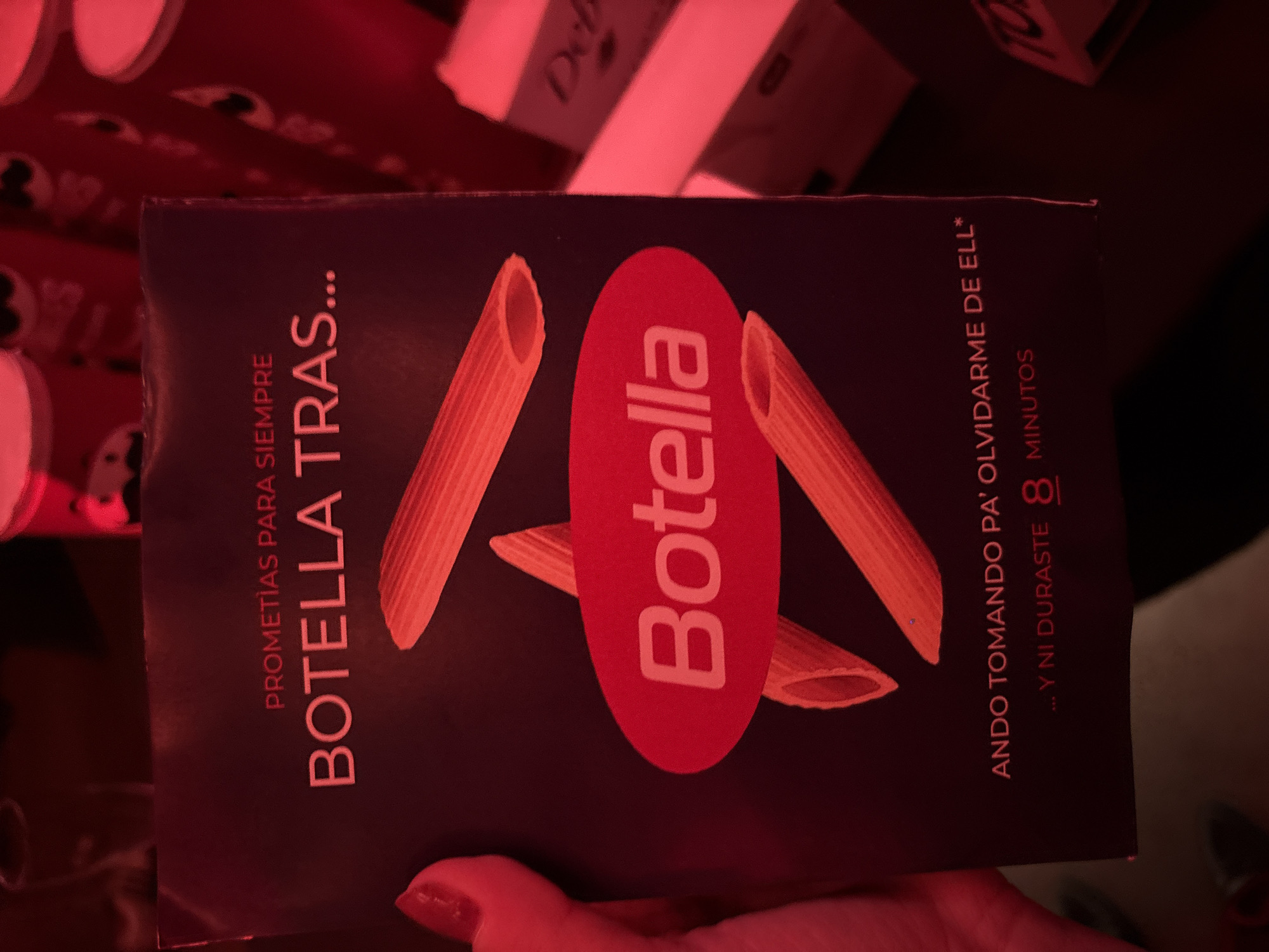 The image shows a vertically oriented package of "Botella" brand pasta, likely in a domestic setting. The package is dark, with images of penne pasta and a red oval with the "Botella" logo. The text on the package is in Spanish and includes the phrase "PROMETIAS PARA SIEMPRE BOTELLA TRAS..." and "ANDO TOMANDO PA' OLVIDARME DE ELL* ... Y NI DURASTE 8 MINUTOS." The package is being held by a hand with a red nail polish. The background is blurry and includes other objects, possibly food or other packages. The lighting is predominantly red, creating a warm tone. There is no information in the image to indicate that the location is Milan, Italy.