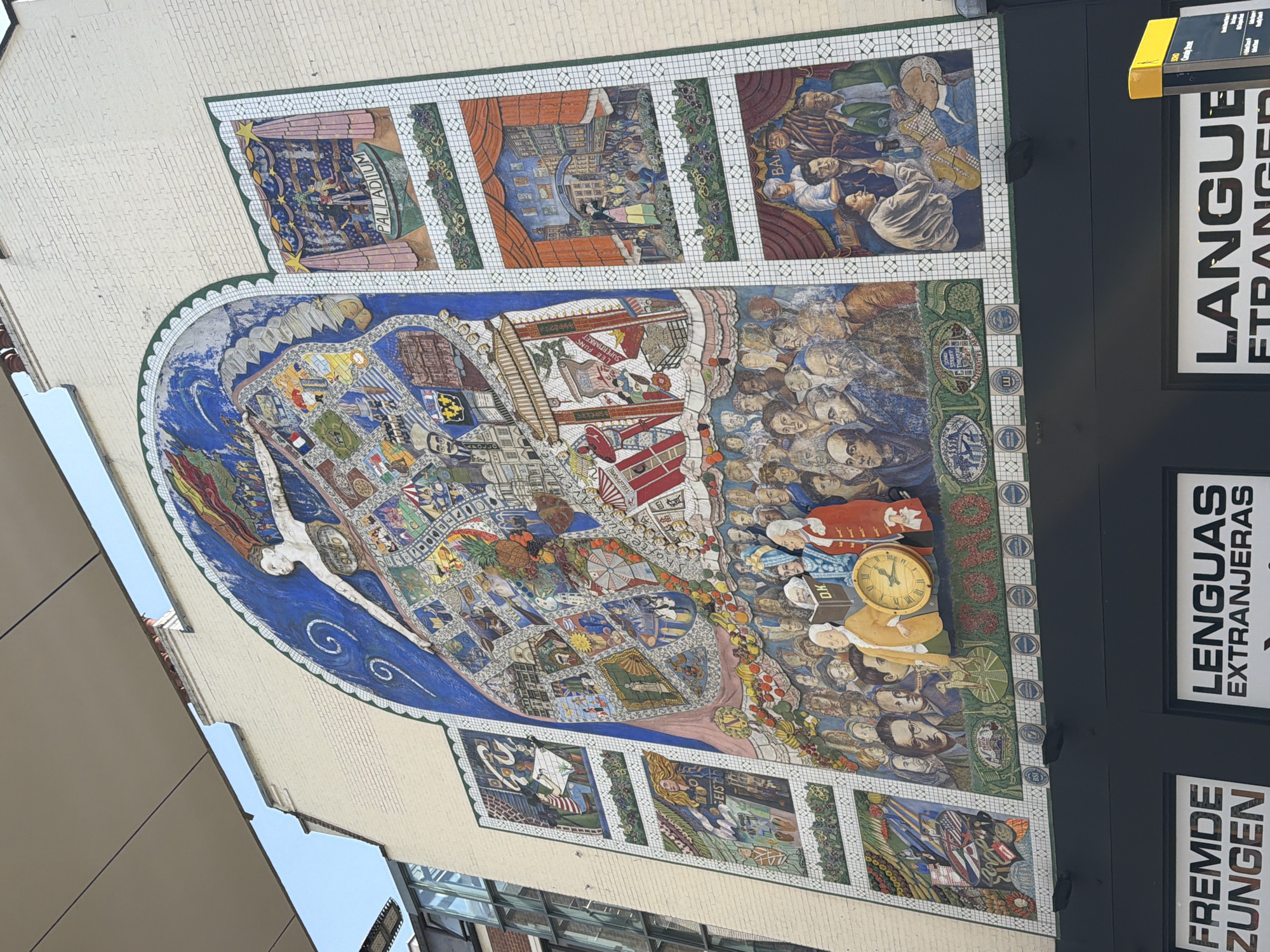The image shows a large mosaic mural on the side of a building. The mural is divided into sections, including a large central image in the shape of an oval, with various scenes and figures depicted within it. The scenes include depictions of buildings, people, and other objects. The artwork is detailed and colorful.
On the right side of the image, there are vertical signs that are likely advertising language classes or translation services, with words such as "LANGUE ETRANGERE," "LENGUAS EXTRANJERAS," and "FREMDE ZUNGEN."
The mural itself includes a depiction of the Palladium theatre. Some businesses referenced in the art include Lee Fung Supermarket.
The building appears to be located in an urban environment, with other buildings visible in the background.
The clock featured in the mural is set to 4:55.
