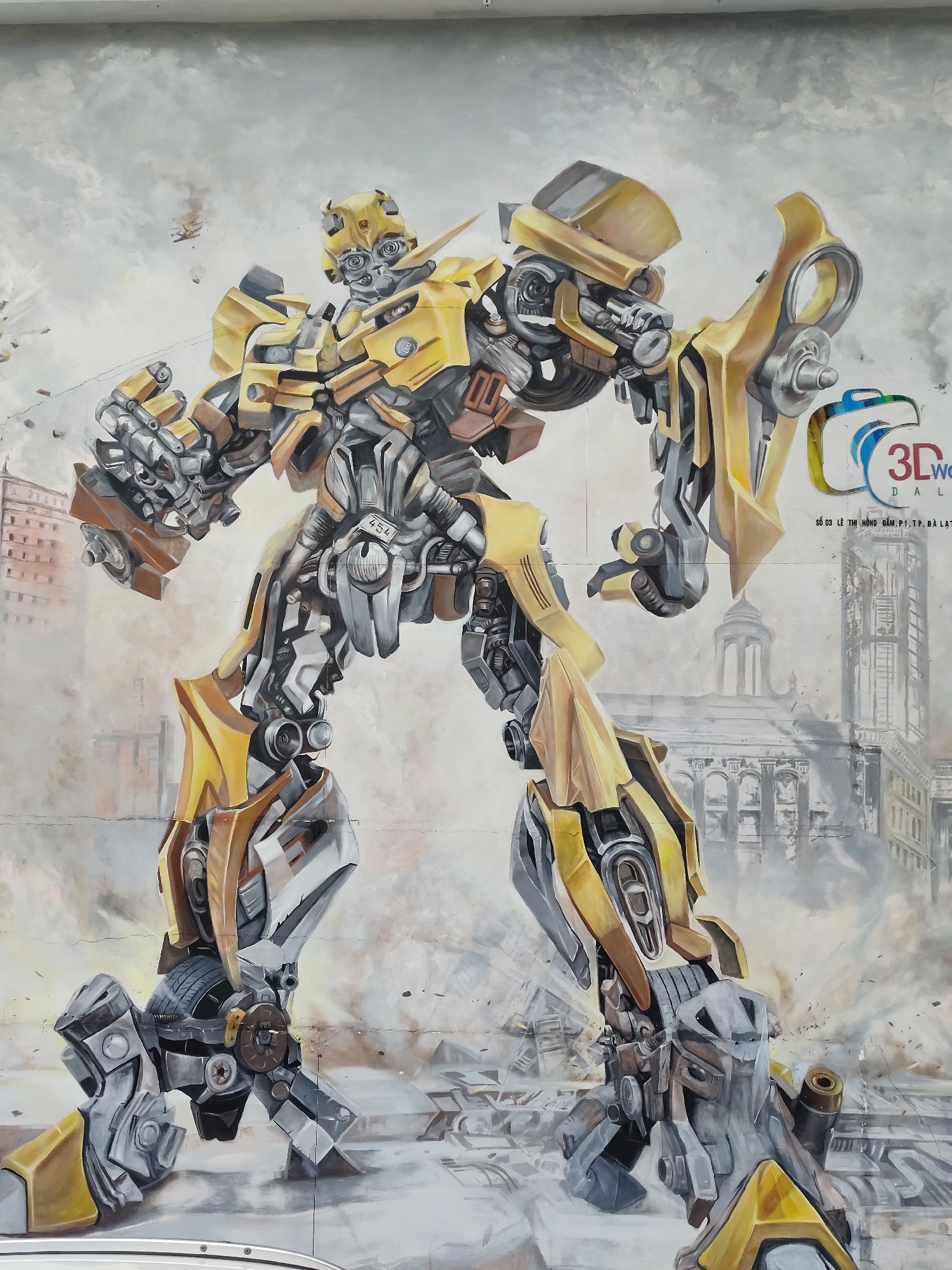 Here is a detailed description of the image:
The image features a large-scale mural of the Transformer Bumblebee painted on an outdoor wall. Bumblebee is depicted in a dynamic pose, with his metallic yellow and gray body prominently displayed. His arms are raised, one clenched in a fist and the other with an open hand.
In the background, a cityscape is painted in grayscale, suggesting a scene of urban decay or war. There is a tall building and a domed structure visible.
To the right side of the mural, there is a logo with "3D world DaLat" written on it, followed by "SỐ 03 LÊ THỊ HỒNG GẤM, P.1, TP. ĐÀ LẠT". This text suggests that the mural is located in Da Lat, Vietnam, specifically at the address: 03 Lê Thị Hồng Gấm, Ward 1, Da Lat City.