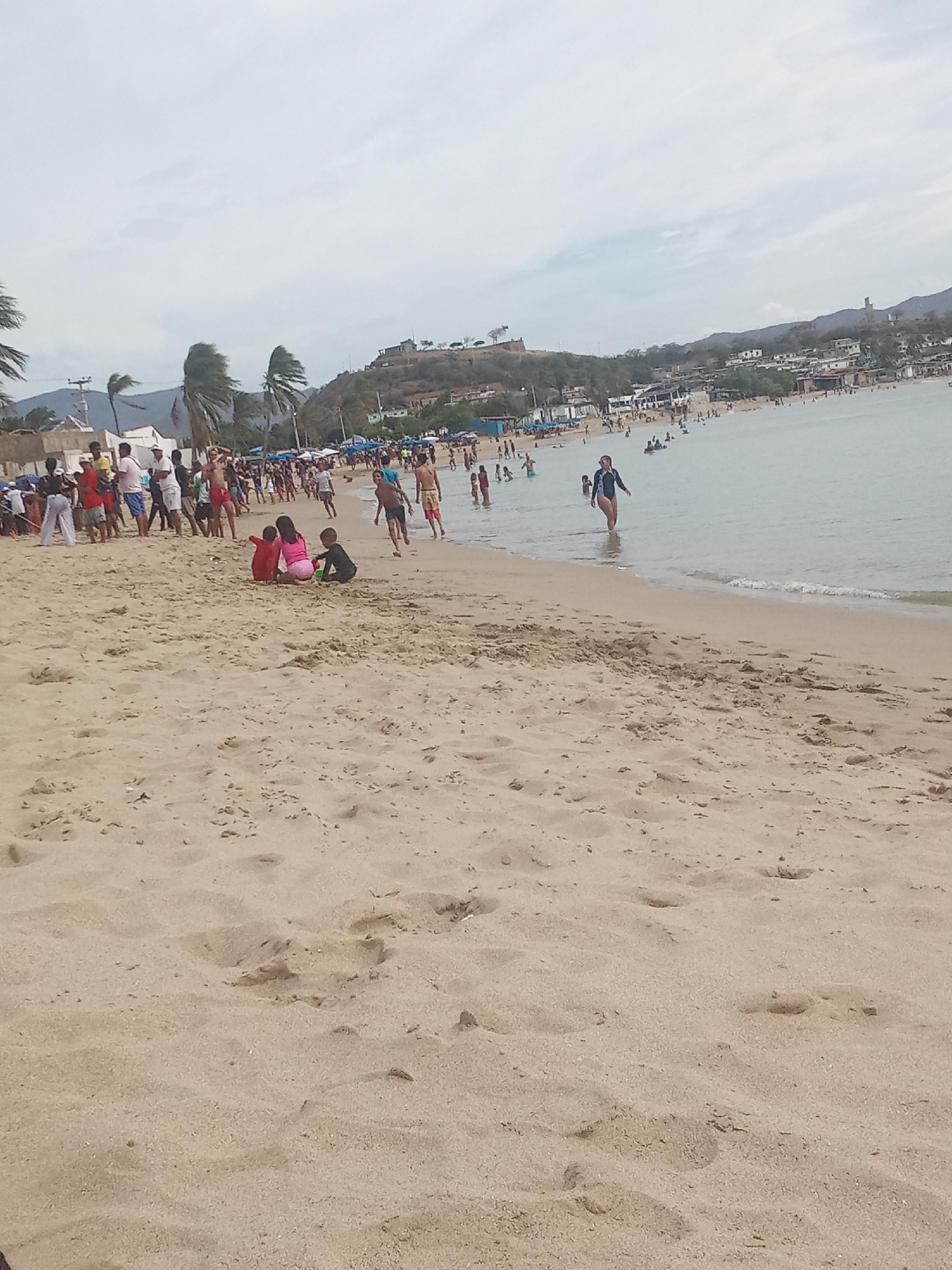 The image shows a crowded beach in Juan Griego, Venezuela. A fort is visible on a hill in the background. In the foreground, there's a sandy beach with people walking, standing, and playing. Some individuals are in the water. The sky is overcast. Palm trees are present on the left side of the image.