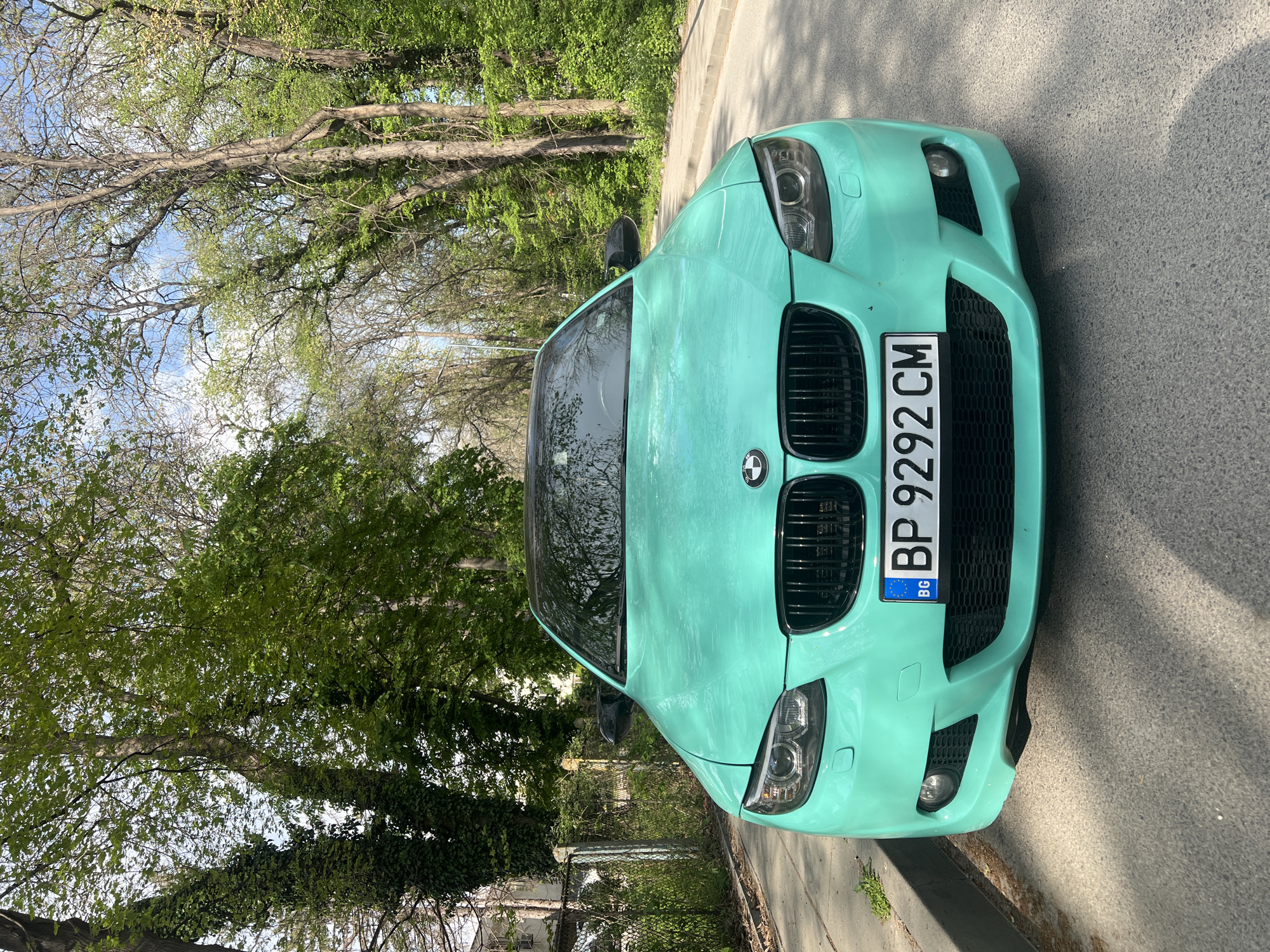 An eye-level shot captures the front of a teal or turquoise BMW E92 3-series coupe, parked on the side of a paved road or path. The car occupies the foreground, facing the viewer directly. Its distinctive black kidney grilles, circular headlights, and lower bumper with fog lights are prominent. The paint or wrap on the car appears slightly textured.
The license plate, centrally positioned on the front bumper, reads "BP 9292 CM" and features the "BG" country code and European Union emblem, indicating its origin from Bulgaria.
The background is dominated by a canopy of trees with dense green foliage, suggesting a lush, natural setting during spring or summer. Some trees have darker, more mature leaves, while others display lighter, fresh green growth. Parts of the sky, a soft blue, are visible through the tree branches. Patches of sunlight filter through the leaves, creating dappled shadows on the ground and parts of the car. No people or additional vehicles are visible, contributing to a serene atmosphere. The scene is well-lit, indicating daytime. Given the context, the location is Plovdiv, Bulgaria.