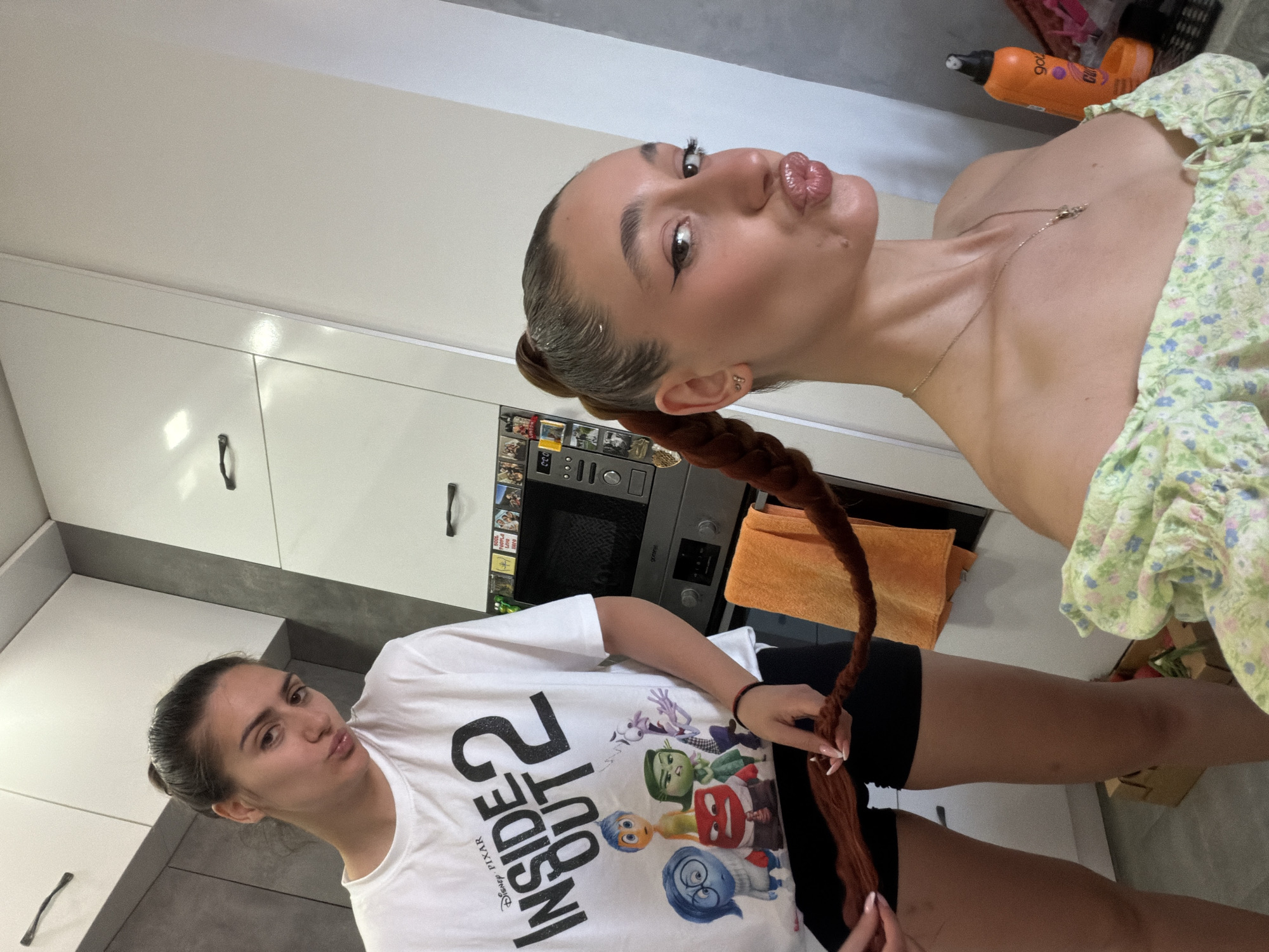 The image shows two young women in a brightly lit, modern kitchen. One woman, in the foreground, is wearing a white t-shirt with a graphic print of characters from Disney Pixar's "Inside Out." She has her hair pulled back and is making a kissing face at the camera. The other woman, in the background, is wearing a light green floral top and has her hair styled in a long braid that extends down her back. She is also making a kissing face. Both women appear to be posing for a selfie. The kitchen has white cabinets and grey countertops, with a microwave and oven visible in the background. There are also some bottles of hair products on a shelf above the counter. The overall mood of the image is playful and casual. The time of day is not specified, but the lighting suggests it is daytime. There is no indication of the weather. The scene appears to be indoors.