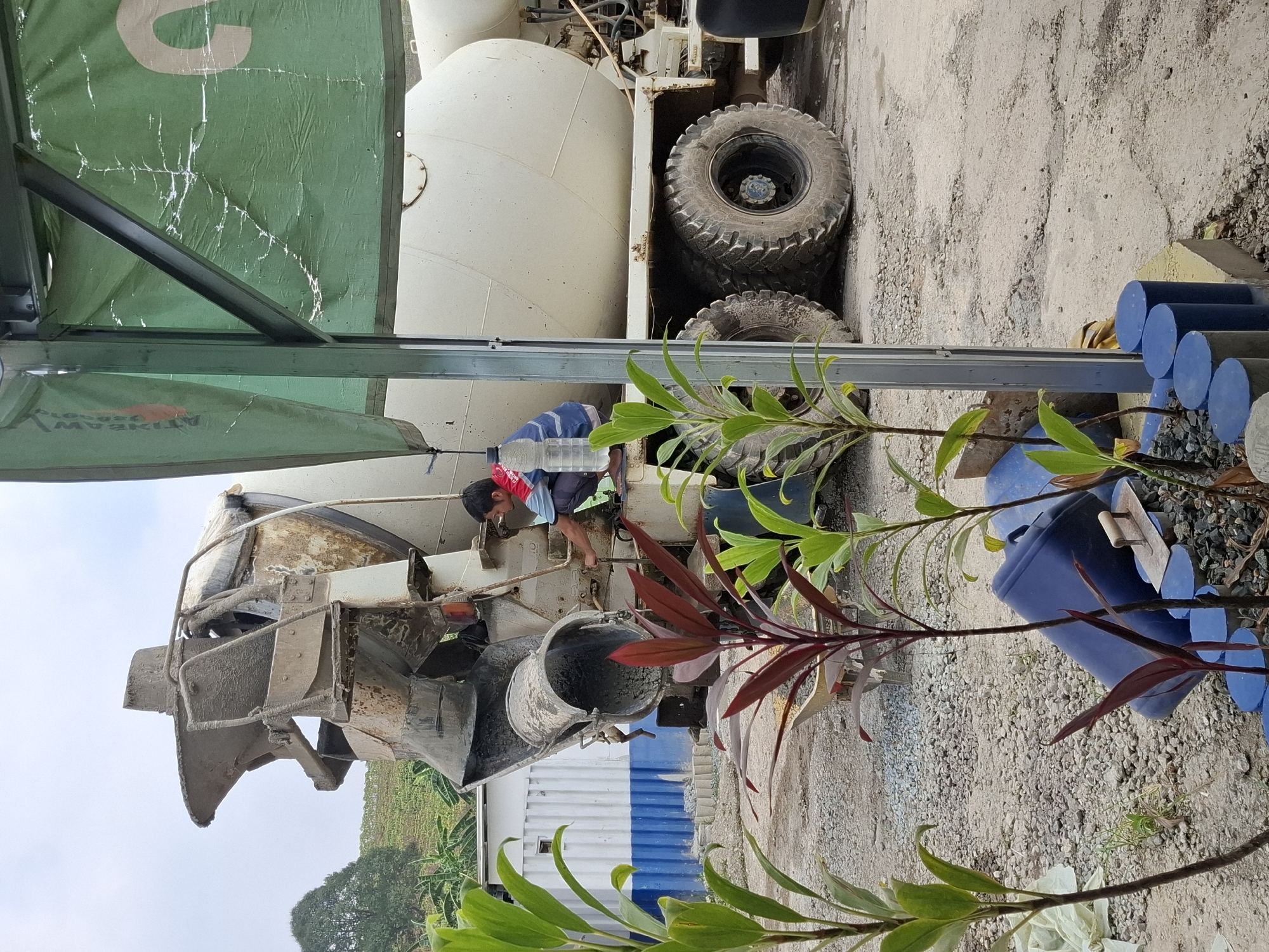 A man in a blue and red shirt is crouched next to a concrete mixer truck in Pamuruyan, Indonesia. He appears to be working on the truck's chute, which is angled downwards, possibly dispensing concrete. The truck is large and appears to be made of white metal. The ground is covered in gravel and dirt, and there are some plants in the foreground. In the background, there is a white and blue building and a green tarp with a large white letter "C" on it. The sky is overcast.