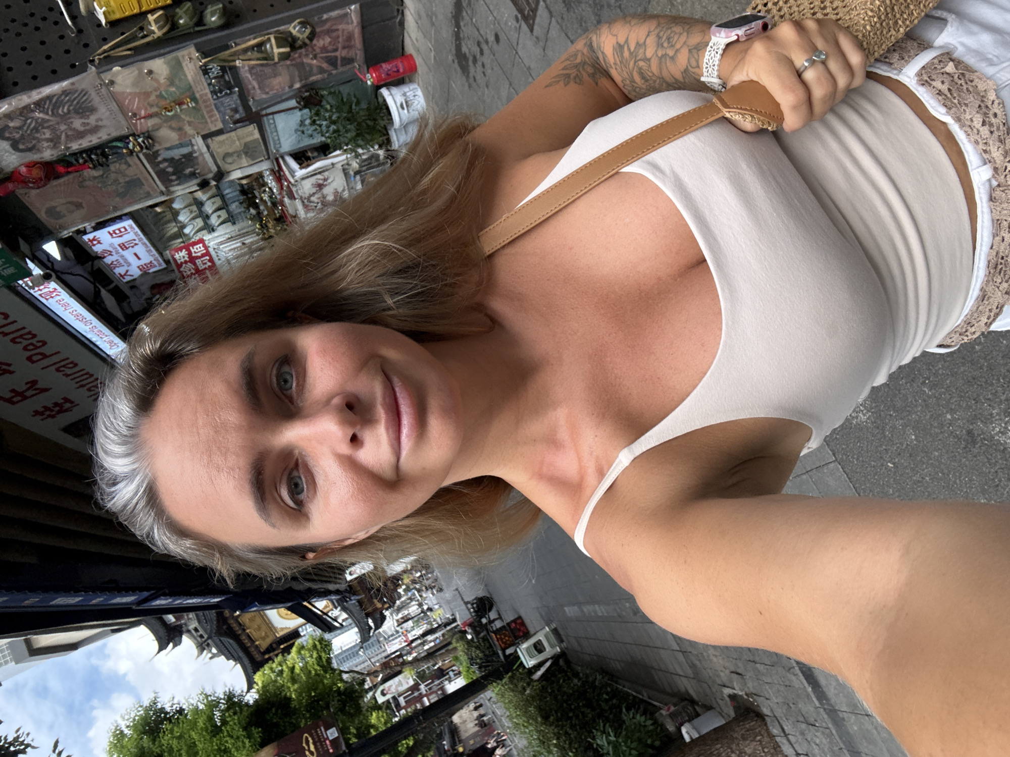 In Shanghai, China, a fair-skinned woman with blonde hair and grey streaks, wearing a white tank top, takes a selfie on a cobblestone street. The street is lined with traditional Chinese architecture on the left and shops with displays of jewelry and art on the right. A brown woven bag hangs from her shoulder, and she is wearing a watch on her left wrist. The background shows a street scene with buildings, trees, and other pedestrians.
