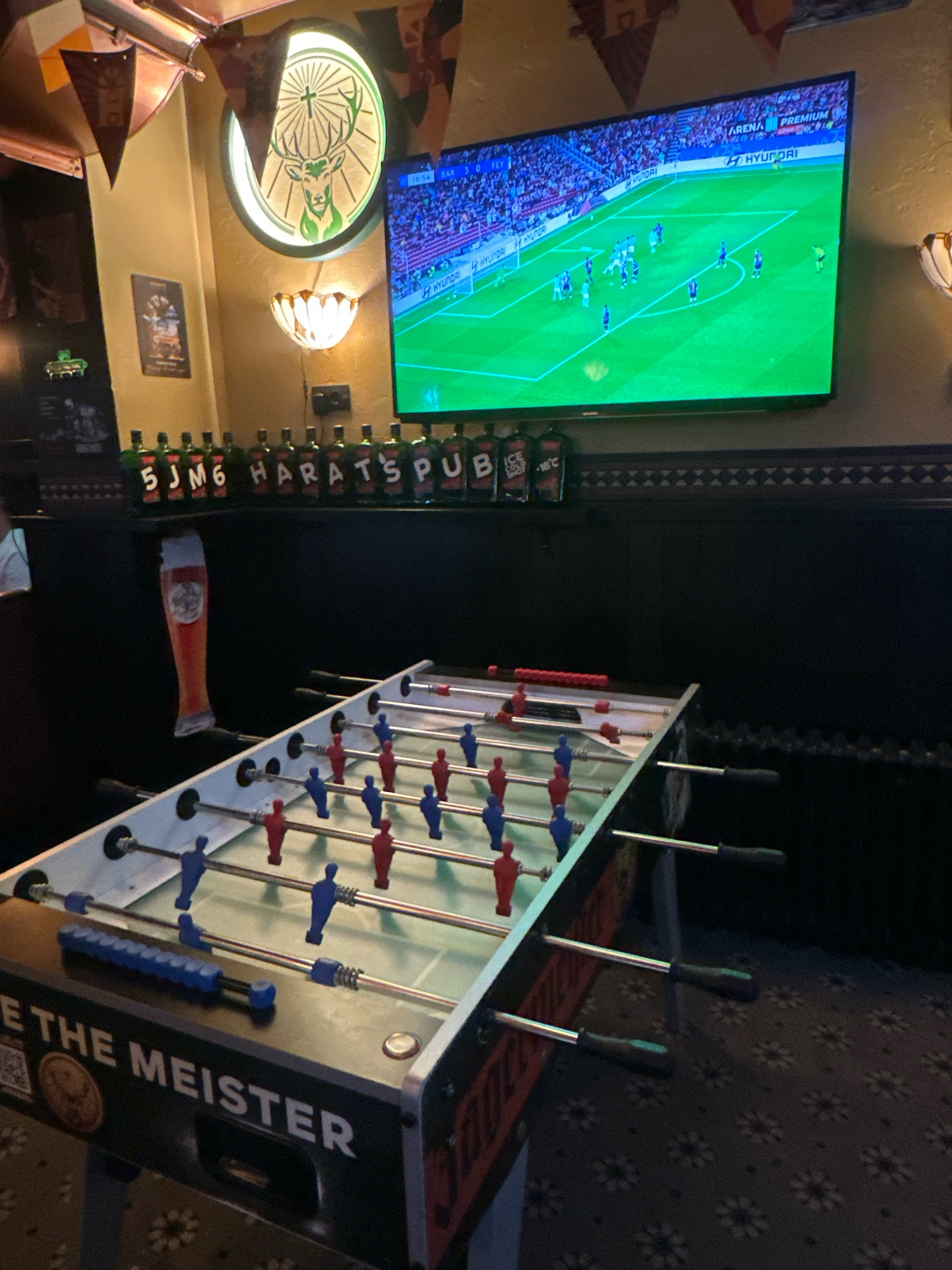 The image depicts an indoor scene, likely a bar or pub, with a foosball table prominently featured in the foreground. The table is white with blue and red player figures on silver rods. A television screen in the background displays a soccer match. Behind the foosball table, a dark wall is visible, adorned with bottles of alcohol arranged on a shelf. To the left of the TV, a circular illuminated sign with a deer logo is mounted on the wall. The overall ambiance suggests a casual entertainment venue. There are no people visible in the image. There is no information about the location being in the city of Belgrade, Serbia.