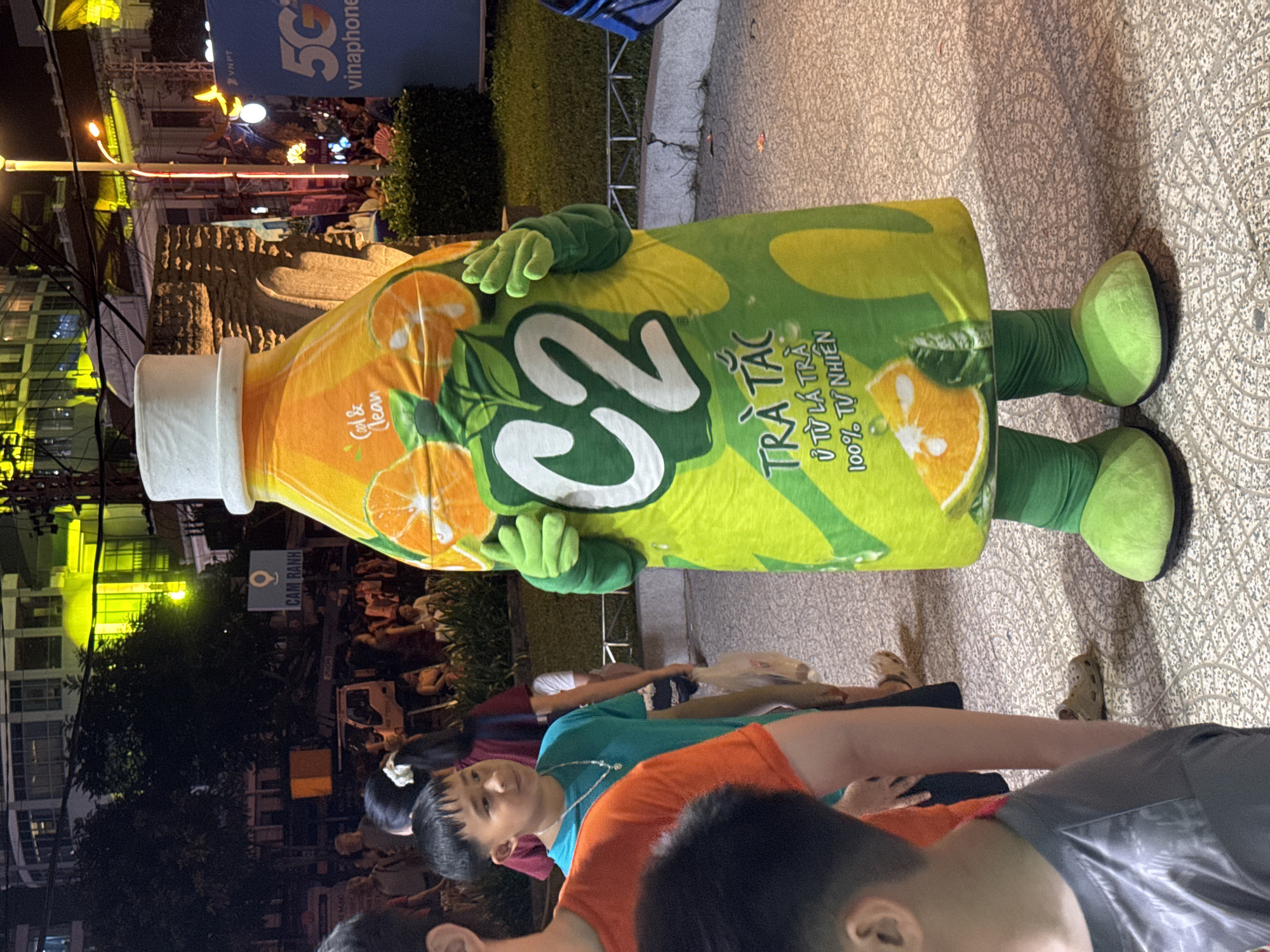 The image captures an outdoor scene at night, likely a public area in Nha Trang, Vietnam, given the Vietnamese text visible on the mascot. A person is wearing a large, mascot costume designed to resemble a bottle of "C2" brand iced tea. The costume is predominantly bright green and yellow, featuring large graphics of oranges and the "C2" logo. The mascot has green gloves and green, oversized shoe covers.
In the foreground, a young boy wearing a teal t-shirt and a silver necklace stands to the left of the mascot. Partially visible to his left is another person in an orange t-shirt. Behind them, other people are milling about in the background. The environment appears to be paved with decorative tiles. In the distance, buildings with bright yellow lighting are visible, along with streetlights and trees. A blue banner with white text, possibly advertising "5G vinaphone," is present in the background to the right. The overall atmosphere suggests a lively, public event or gathering.