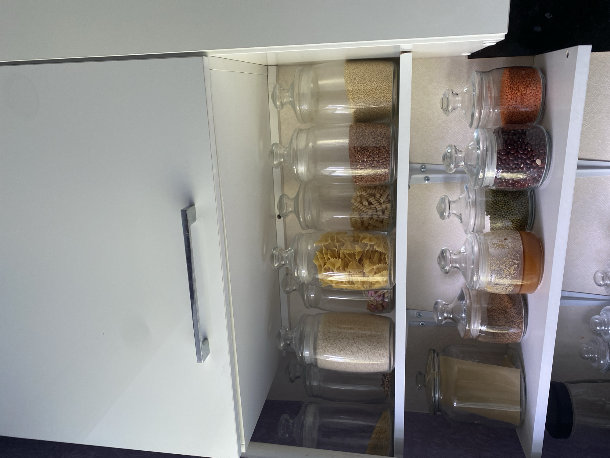 This is a medium shot, eye-level, interior photograph of a kitchen cupboard in Lomonosov, Russia. The cupboard is white with a brushed metal handle on the upper cabinet door. The shelves are white. The top shelf contains seven glass jars filled with dry goods. From left to right, the jars contain: brown granules, small dark beans, white rice, pinkish-red dried flowers, yellow bow-tie pasta, small speckled beans, and yellow couscous. The second shelf contains four glass jars. From left to right, the jars contain: a large jar with a brown paper insert, a jar with instant coffee granules, a jar with a golden liquid, and a jar with green lentils. To the right of the green lentils, three more jars are visible containing red kidney beans, and orange lentils. The lower shelves contain more glass jars with lids. The overall environment is clean and organized.