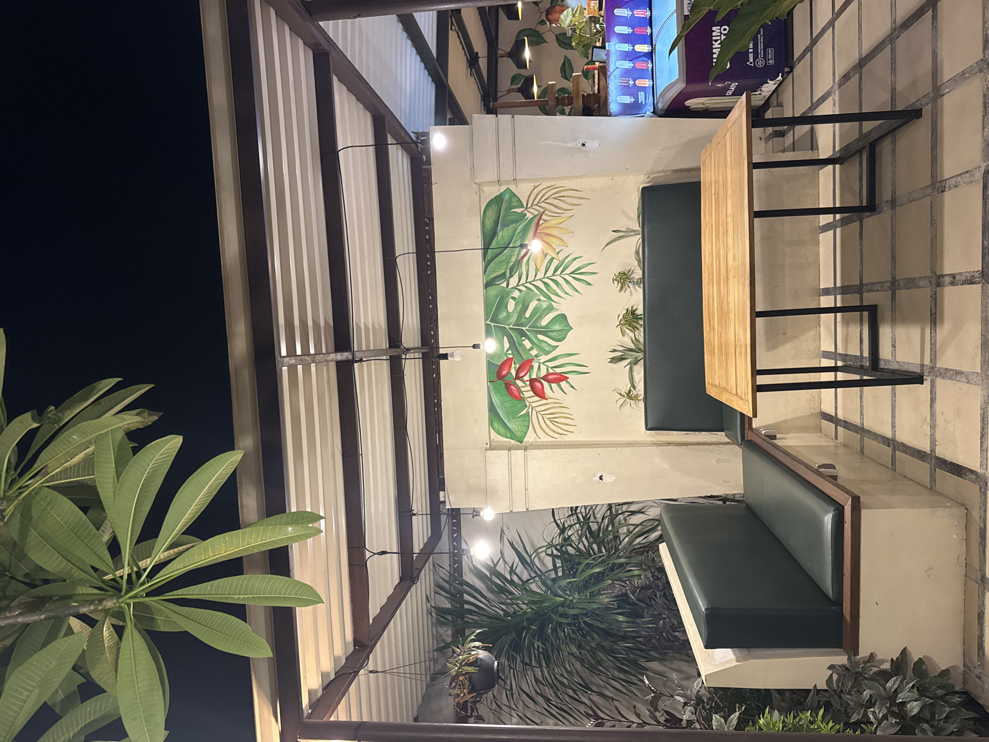 An outdoor seating area in Kuta Selatan, Indonesia, features a wooden table with black metal legs, surrounded by a built-in L-shaped bench upholstered in dark green leather. A tropical-themed mural adorns the back wall, depicting large green leaves and red flowers. The area is covered by a corrugated metal roof supported by a metal frame, with hanging plants and bare lightbulbs visible. In the background, an ice cream freezer with colorful popsicles is visible, along with additional seating and foliage. The ground is paved with square tiles.