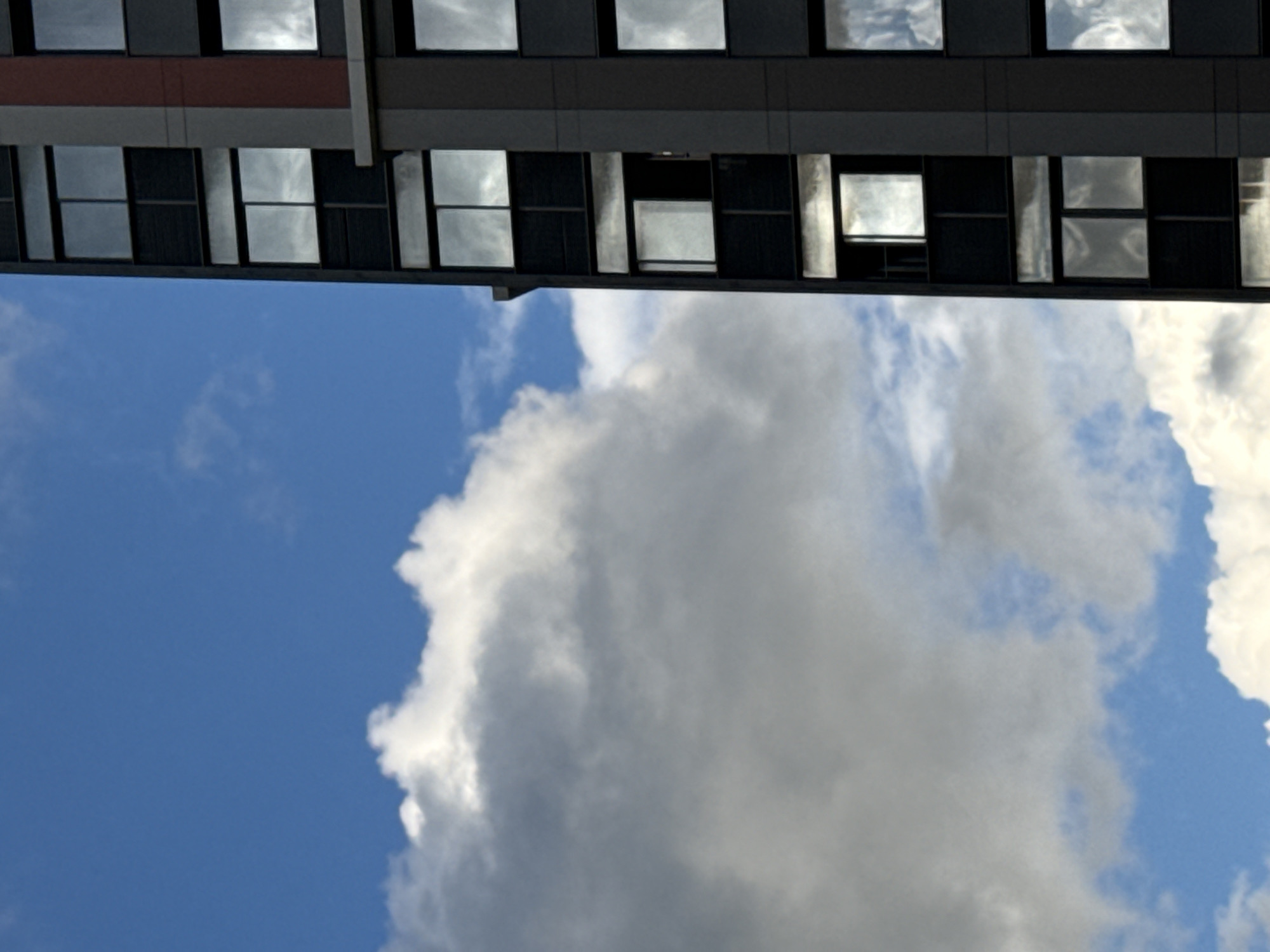 This is a vertical, medium-shot, eye-level photograph taken on a sunny day with a partly cloudy sky, featuring a modern building with dark gray and red accents, and large reflective windows. The building occupies the right side of the frame, its facade showing a grid of windows, many of which reflect the white, billowy clouds and the bright blue sky. A notable feature is a vertical red stripe running down the side of the building. The left side of the image is dominated by a large, bright white cloud formation against a clear blue sky. The scene appears to be an urban environment, though no specific landmarks or people are visible, making it impossible to determine the exact location within Moscow, Russia.