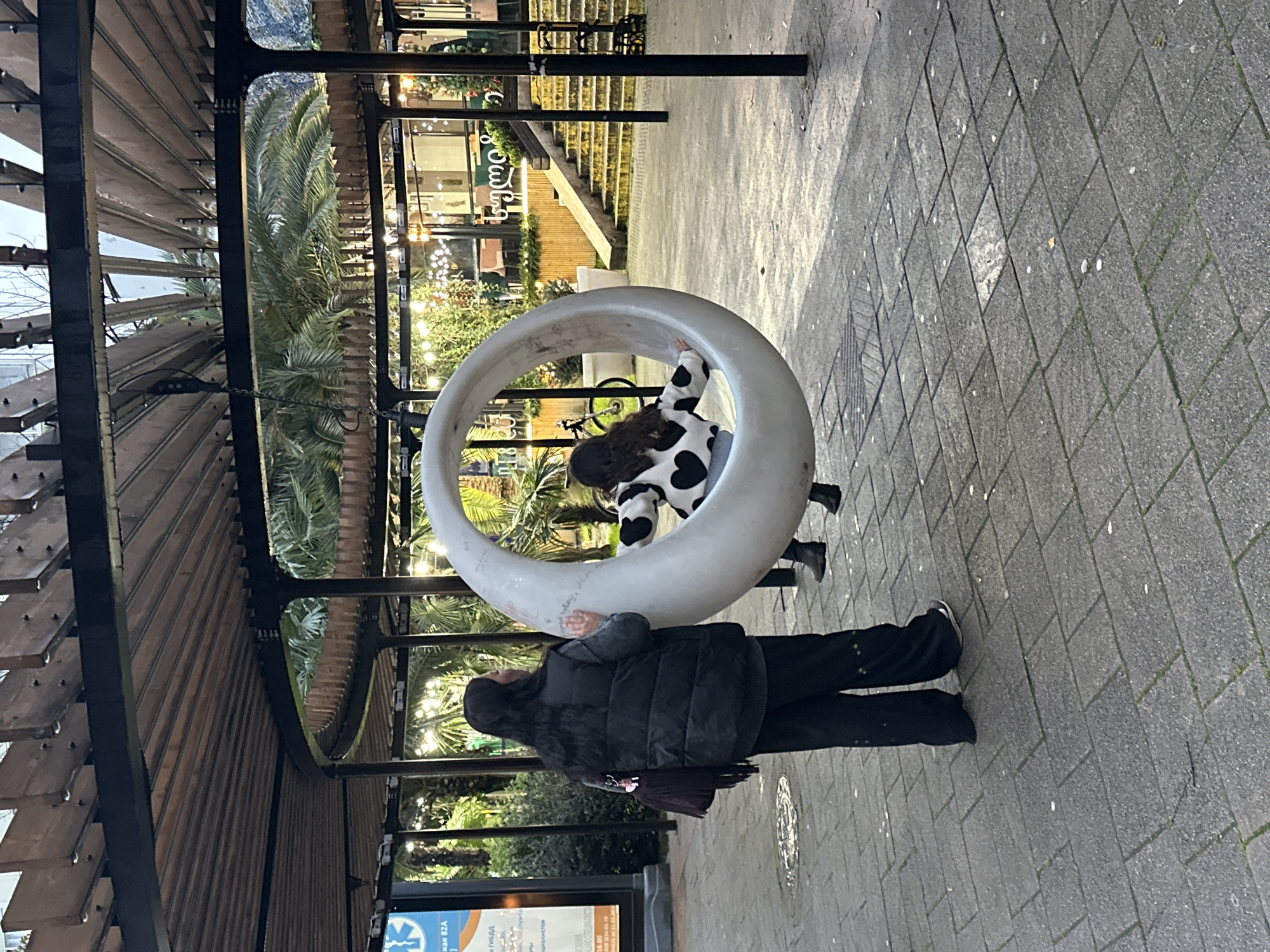 In Sochi, Russia, an outdoor scene on a paved walkway features a large crescent moon-shaped swing. A child wearing a black and white cow-print jacket is inside the swing. A woman in a black puffer jacket and black pants is standing near the swing. A pergola structure with wooden slats is visible on the left side of the frame. Palm trees and buildings with lights are in the background. A sign with text is in the lower-left corner.