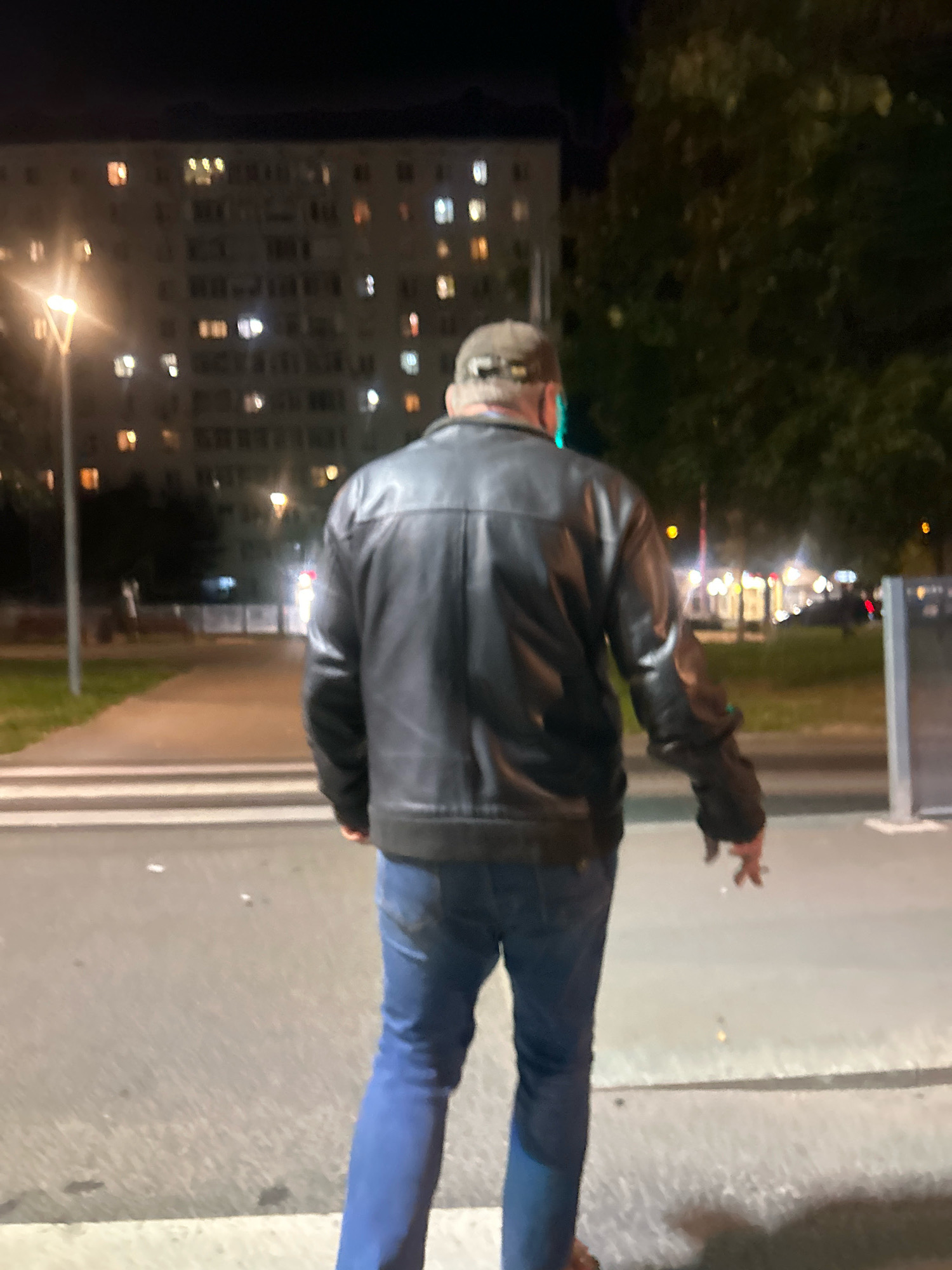 A man in a black leather jacket and blue jeans is standing on a crosswalk at night. He is facing away from the camera and is holding a cigarette in his right hand. In the background, there is a large apartment building with illuminated windows, streetlights, trees, and other buildings. The scene appears to be in an urban environment. The location is identified as Moscow, Russia.