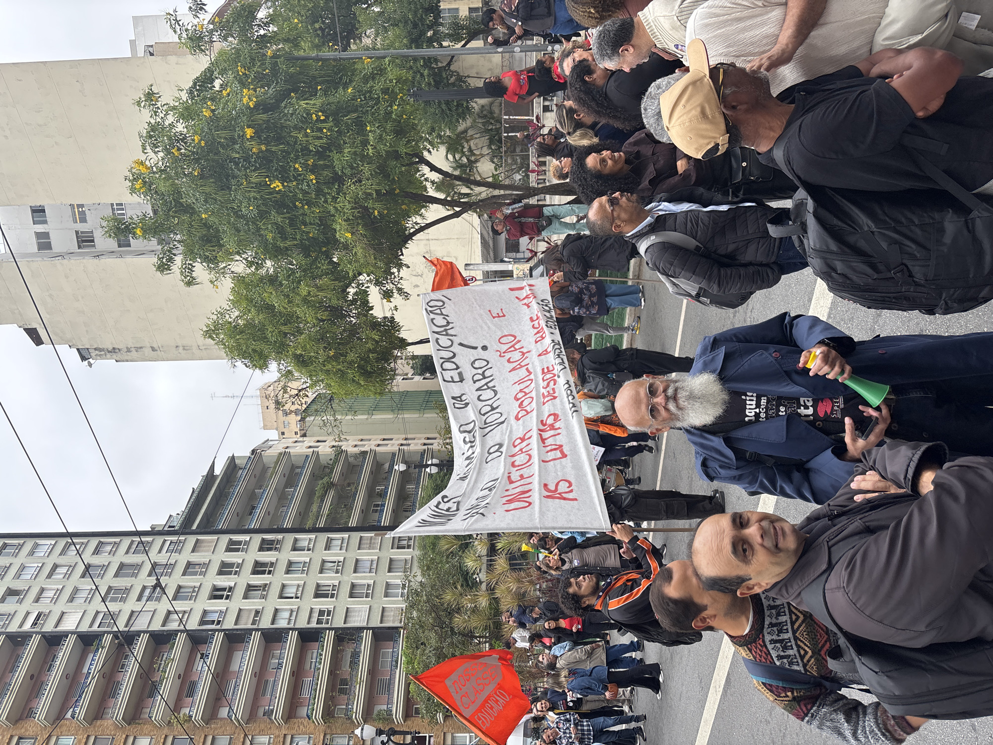 The image depicts a street-level view of a protest in an urban setting, likely São Paulo, Brazil, given the building architecture and the Portuguese text on the banners. A crowd of people is gathered on a street, some holding banners. The banner visible reads, "Unificar Populacão e as Lutas desde a Base." Another sign reads "Nossa classe Educação". Many protesters can be seen in the image. Buildings line the sides of the street, and a tree with yellow flowers is visible in the foreground. A man in the foreground looks directly at the camera.