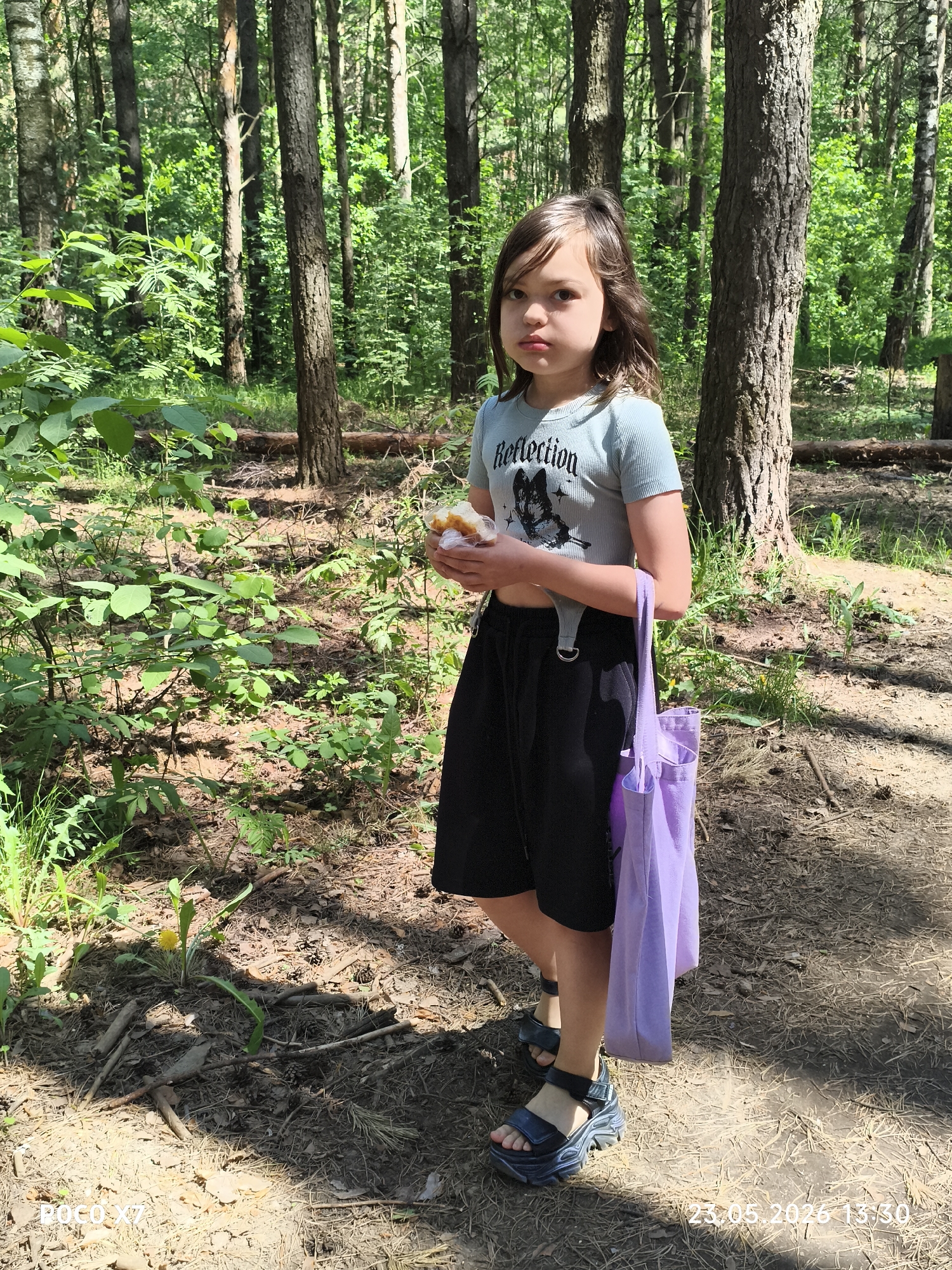 The image shows a young girl standing in a forest in Chaikovsky, Russia. She is wearing a light blue t-shirt with the word "Reflection" printed on it, black shorts with suspenders, and chunky black sandals. She is holding a wrapped food item in her hands and has a lavender tote bag hanging from her shoulder. The background is a dense forest with tall trees and green foliage. The ground is covered in leaves, pine needles, and some small plants. The timestamp in the lower right corner indicates the photo was taken on 23.05.2026 at 13:30.