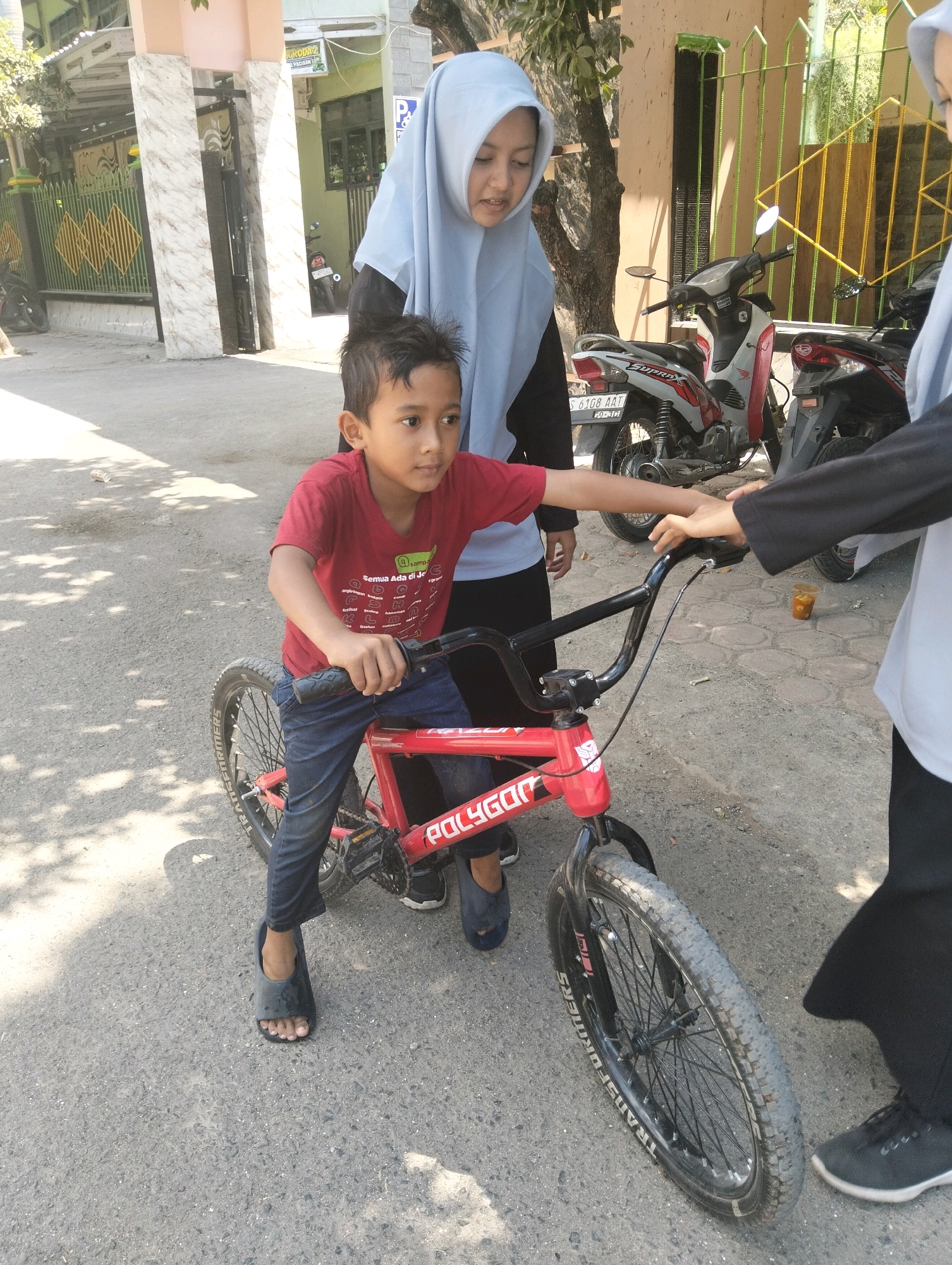 The image depicts a street scene in Paciran, Indonesia, with a young boy seated on a red bicycle. He is wearing a red t-shirt and dark blue jeans. Two individuals, likely female, are also present. One is wearing a light blue hijab and a dark long-sleeved shirt, and the other is wearing a light grey long-sleeved top and black pants. The boy is holding onto the handlebars of the bicycle, and one of the women's hands is near the handlebars, suggesting an interaction. In the background, two motorcycles are parked, and a building with a green wall and a yellow gate is visible. The ground is paved with asphalt and there are shadows from trees. A small plastic cup with liquid is on the ground.