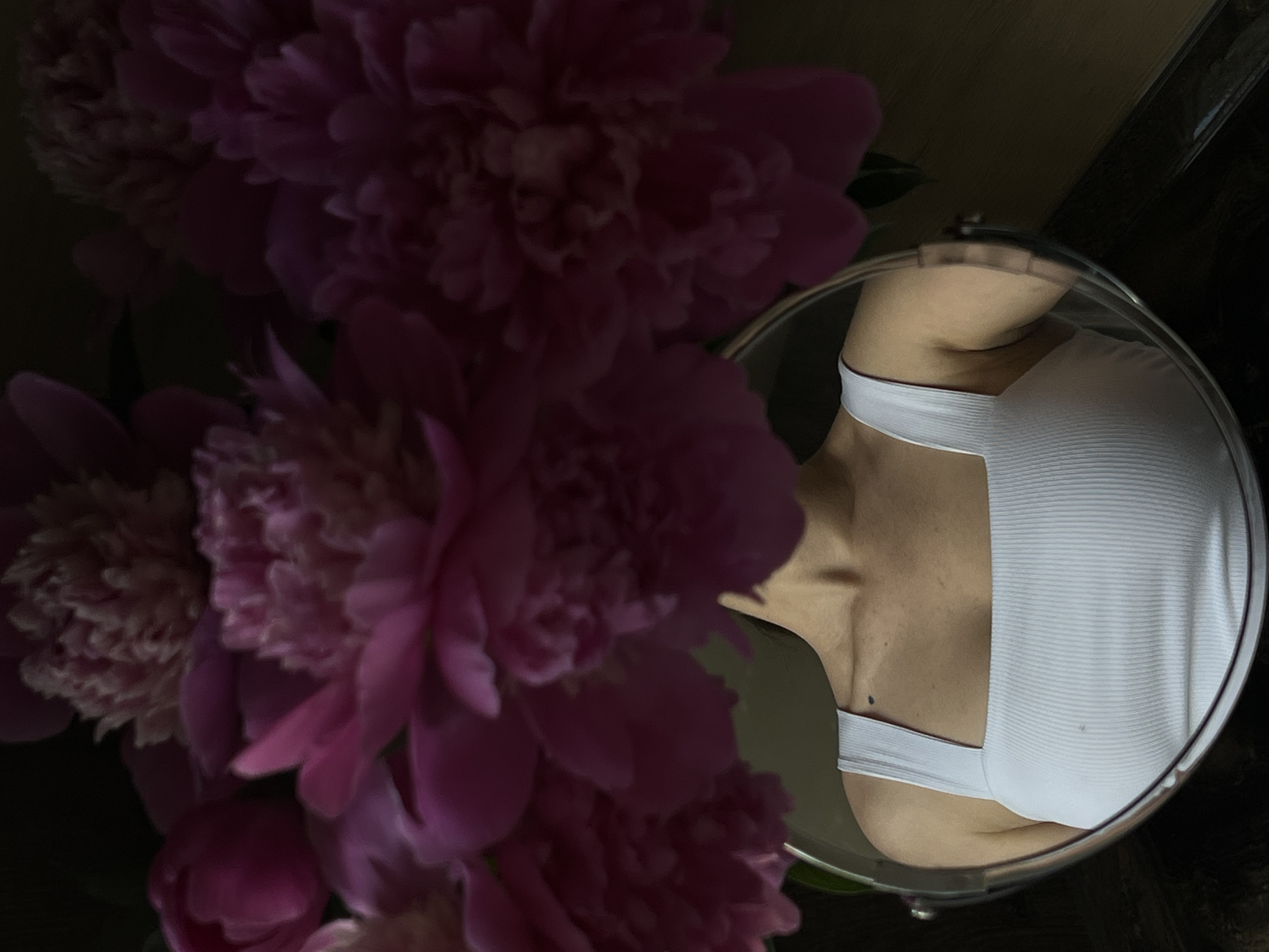A close-up shot focuses on a mirror reflecting the upper body of a person wearing a white ribbed tank top. The tank top has square neckline and thin straps. A dark mole is visible on the left collarbone area. The mirror is round with a thin metallic rim and is positioned in front of a dark, textured background. Above and behind the mirror, a bouquet of large, pink peonies is prominently displayed, with their full petals filling much of the upper frame. The lighting is dim, creating deep shadows and highlighting the textures of the flowers and the fabric of the tank top. The scene is likely set indoors, possibly in an apartment or a studio in Ternopil, Ukraine.