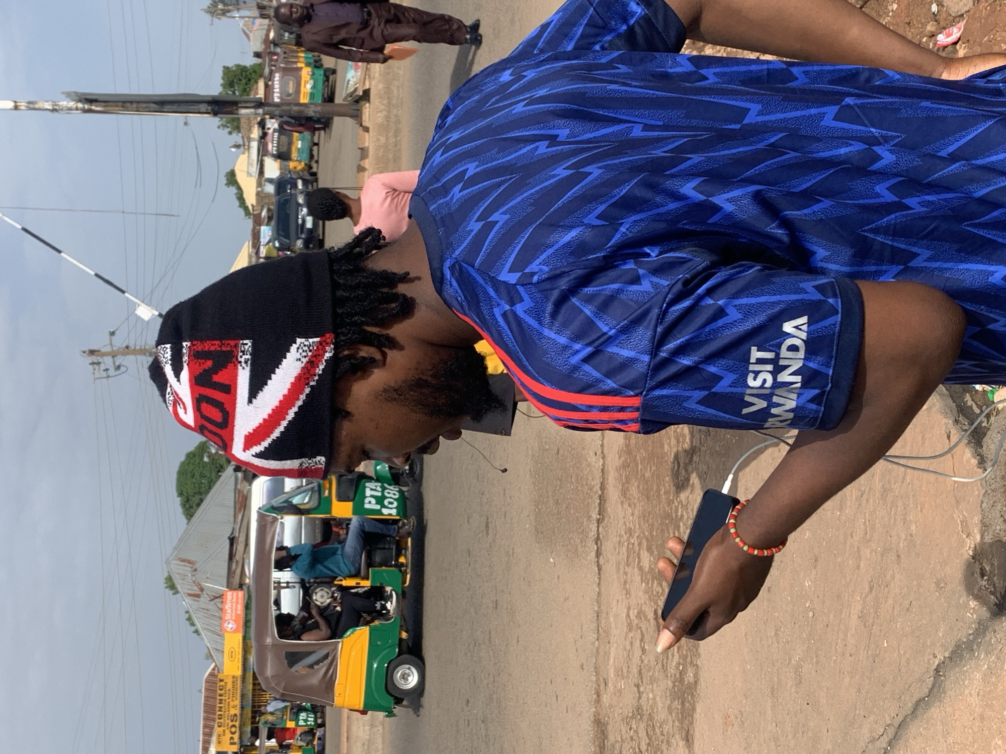In Gura Top, Nigeria, a young man wearing a black beanie with a Union Jack design and a blue patterned t-shirt is standing on a dirt road, looking at his phone, which is plugged into a power bank attached to his wrist with a beaded bracelet. Behind him, several motorized rickshaws, also known as 'okadas' or 'keke napep,' are stopped or moving, indicating a busy street scene. One rickshaw has passengers, and another has advertisements for goods and services. In the background, various buildings and structures, including a tin-roofed shop with yellow signage, line the road. The sky is overcast, and the overall environment suggests a typical day in a bustling Nigerian town.