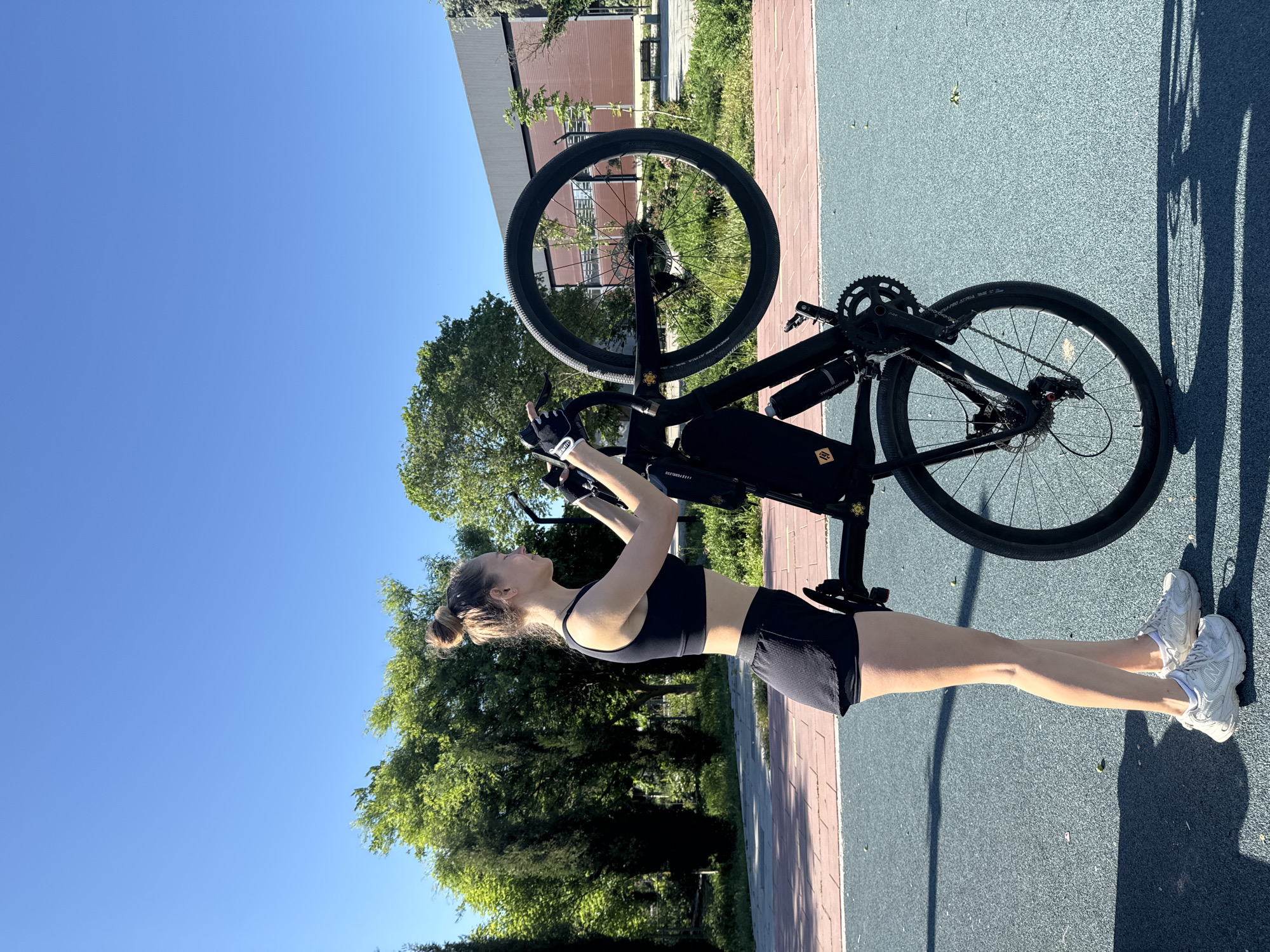 A young woman in athletic attire is holding a black bicycle above her head outdoors on what appears to be a multi-use path. She wears a black sports bra and shorts, along with white sneakers and cycling gloves. The bicycle is also black, with a water bottle holder and a bag attached to the frame. The background consists of green trees, a building, and a clear blue sky. The location appears to be a park or recreational area, potentially within the city of Мысовское сельское поселение, Ukraine, based on the user's information.