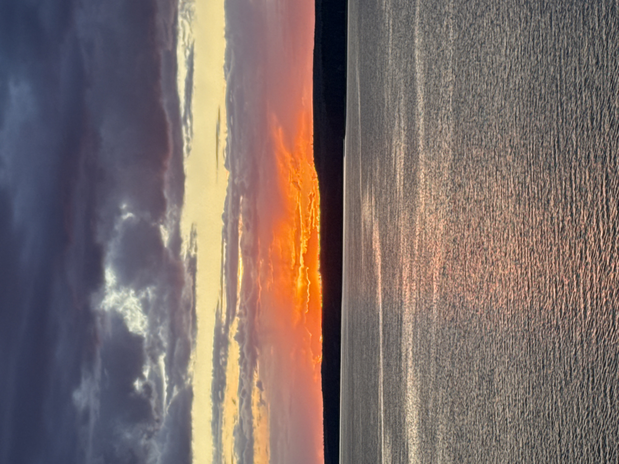 The image depicts a landscape at sunset or sunrise, likely near a body of water. The left side of the image features a sky with a gradient of colors, ranging from dark, stormy grays and purples to bright yellows, oranges, and pinks near the center. These colors suggest the presence of clouds.
On the right side is what appears to be a body of water, possibly a lake or a river, with the same colors reflecting off the sky. The water has a textured surface, indicating some wind or movement. A dark band separates the sky and the water, suggesting a shoreline or horizon.
Given the context provided - that the image is taken in the city of Votkinsk, Russia - the body of water is probably the Votkinsk Reservoir, one of the largest artificial lakes in the region. This detail is critical for precise identification. The photograph captures a moment of natural beauty, focusing on the vibrant colors of the sky and their reflection on the water.