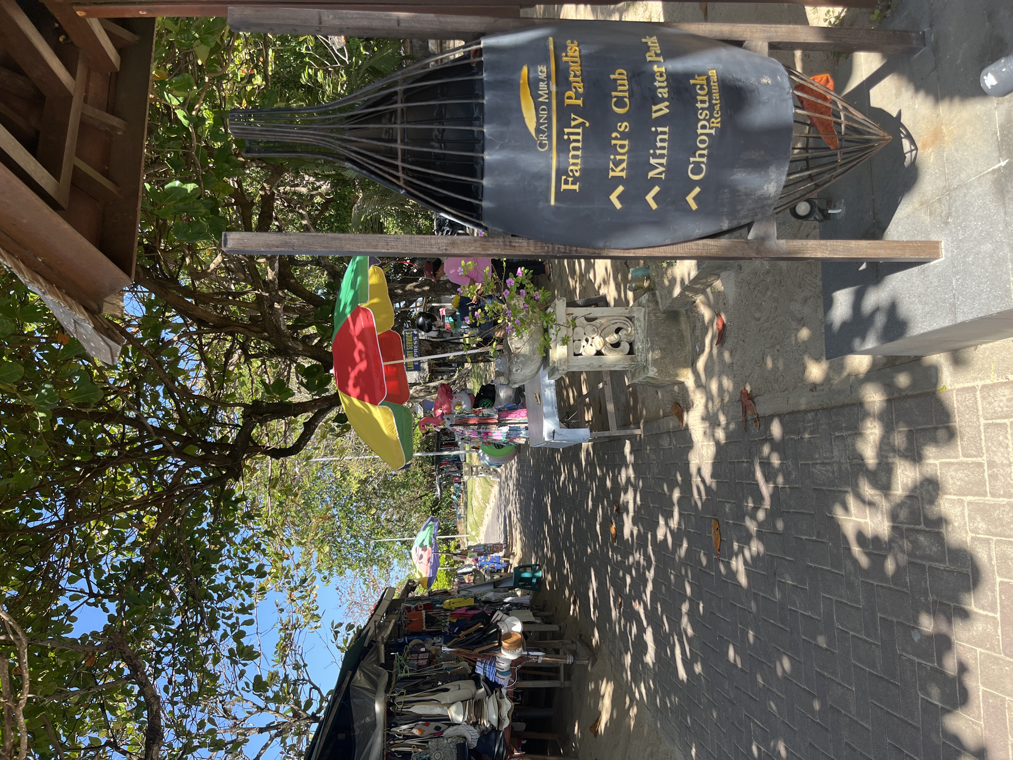The image captures a sun-dappled pathway lined with market stalls and trees, suggesting a location in a tropical resort area. The pathway itself is paved with interlocking bricks, casting dappled shadows from the overhead foliage. On the left, a stall displays an assortment of merchandise, including hats, bags, and clothing, hanging from its structure. Colorful umbrellas, a red, yellow, and green one, and another with a floral pattern, provide shade over some of the stalls.
Further down the pathway, more stalls and vendors are visible, with people browsing or working. A woman in a pink shirt is seen near a stall with flowers. In the foreground on the right, a large, black, tear-drop shaped sign is mounted on a wooden frame. The sign, with the logo "GRAND MIRAGE" at the top, lists amenities such as "Family Paradise," "Kid's Club," "Mini Water Park," and "Chopstick Restaurant." Below the sign, a metal cage-like structure contains some orange material. The overall environment appears to be outdoors, likely a pedestrian walkway in a tourist-frequented area. The presence of tropical trees, beachwear items for sale, and the general ambiance points towards a location in Kuta Selatan, Indonesia, known for its beaches and resorts.