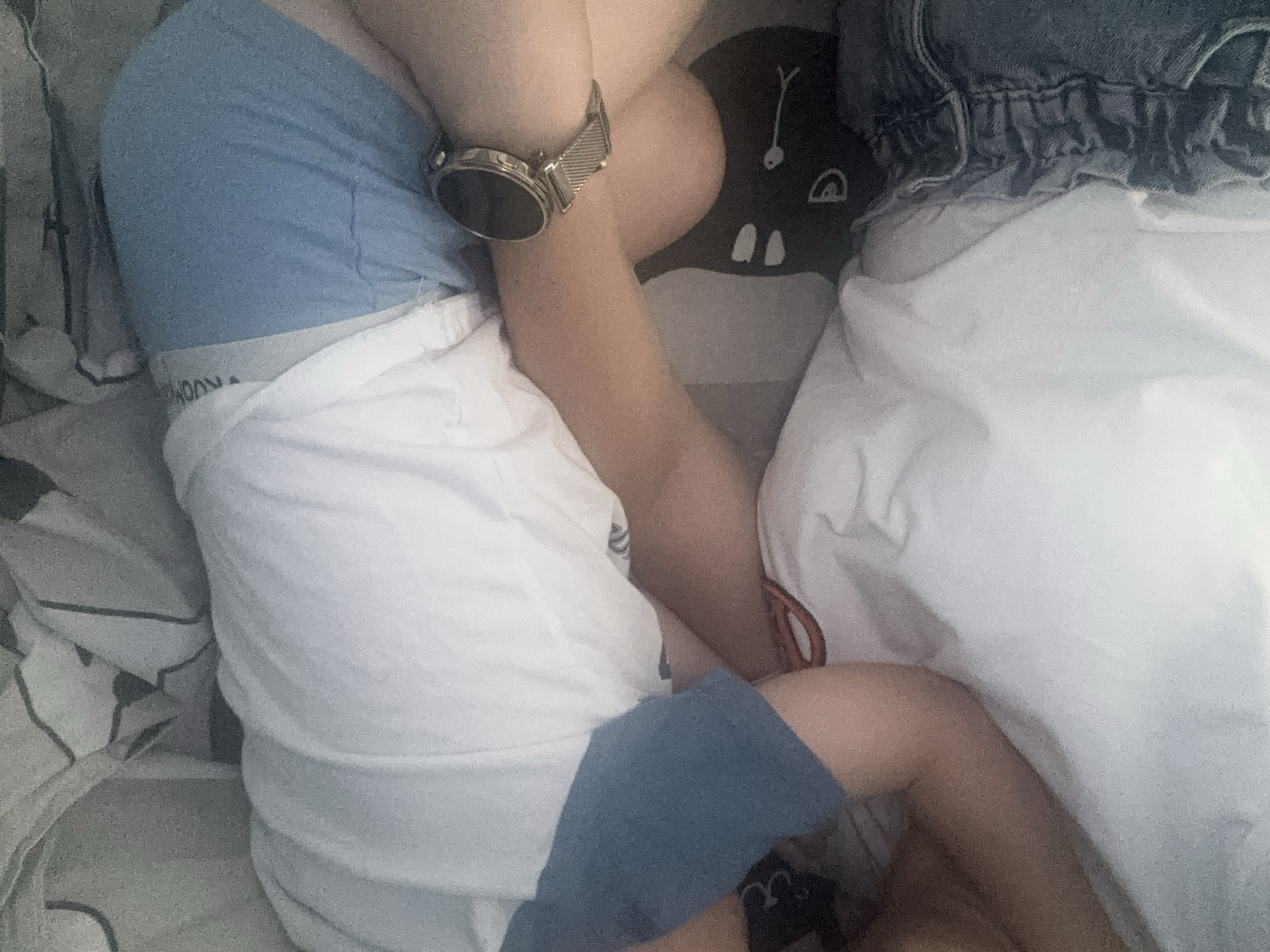 The image shows a close-up of a person lying down, with their legs bent and crossed. They are wearing blue shorts over white underwear, and a white t-shirt. A silver smartwatch with a mesh band is visible on their left wrist. To the right of the person is a white duvet or blanket, and above that, a pair of denim jeans. To the left of the person are patterned sheets. The background includes a black mat with a cartoonish white face design. The lighting is dim, suggesting it might be nighttime or indoors with artificial lighting. The image quality is slightly grainy. There is no discernible text, other than what appears to be a logo or text on the white underwear, which is partially obscured and illegible. The overall impression is of someone resting or relaxing in a domestic setting. The location context of Burgas, Bulgaria, does not offer any specific visual cues within the image.
