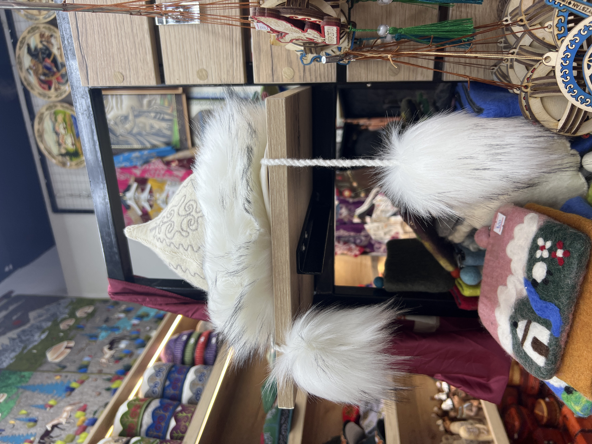 This image depicts a close-up, eye-level view of a market stall in Bishkek City, Kyrgyzstan. The foreground is dominated by a white, fluffy hat with a fur trim, possibly a traditional Kyrgyz hat, and two large white pom-poms hanging from strings attached to the hat. Below the hat, on a wooden shelf, is a felt item with an embroidered landscape of a house, water, and snowy mountains. To the right of the hat, various wooden ornaments with intricate designs, some featuring birds and Cyrillic text, hang from strings. The background reveals shelves filled with colorful hats, tapestries with traditional motifs depicting horses and yurts, and decorative plates. The overall impression is of a vibrant souvenir stall showcasing Kyrgyz craftsmanship.