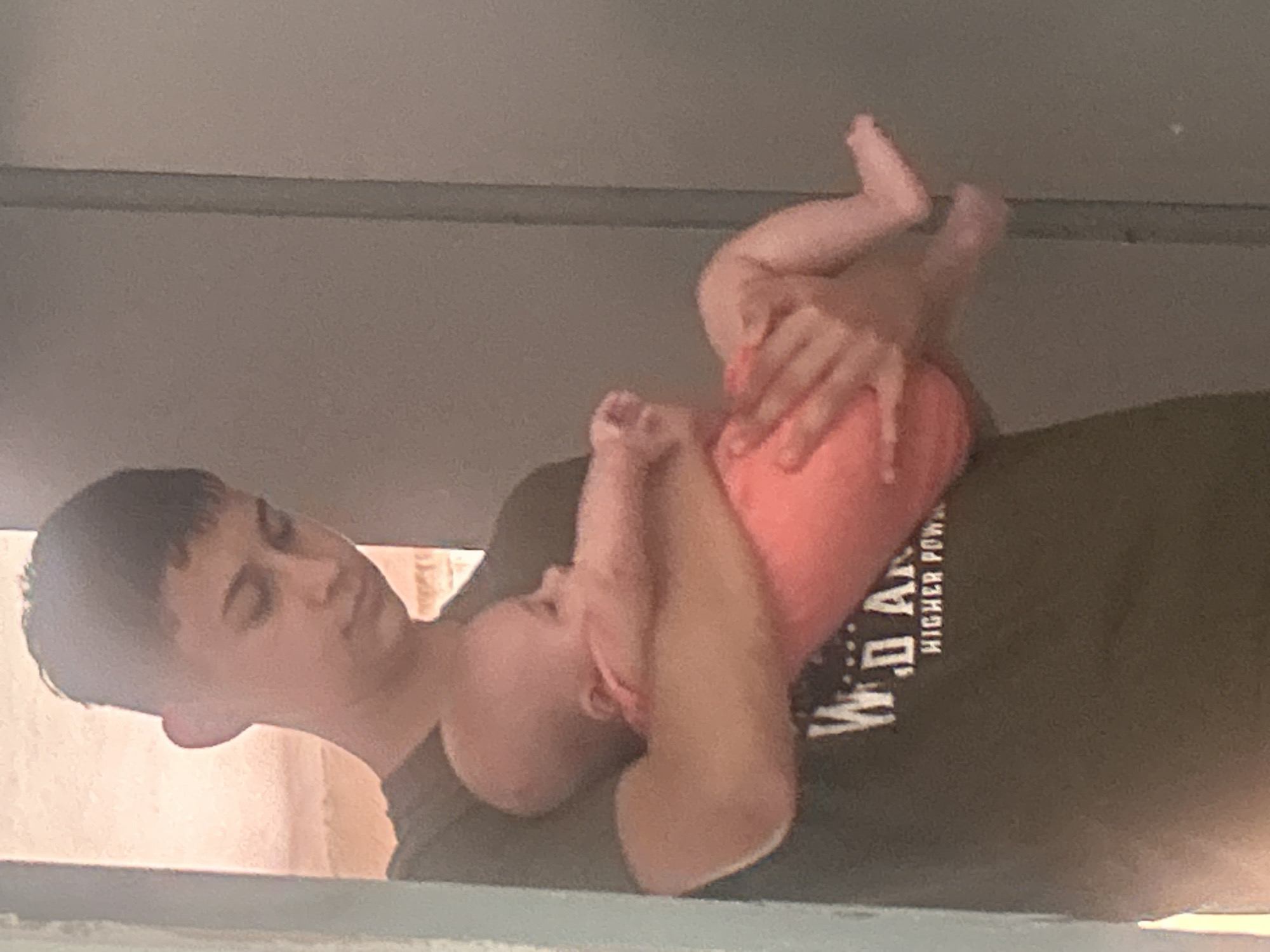 The image shows a young man holding a baby. The man is wearing a dark t-shirt with white text on it. The baby is wearing a pink outfit and is being held in the man's arms. The background appears to be an indoor setting with a doorway or wall on the right side. The image does not provide any information about the location in Govedare, Bulgaria, or any other specific geographic context.