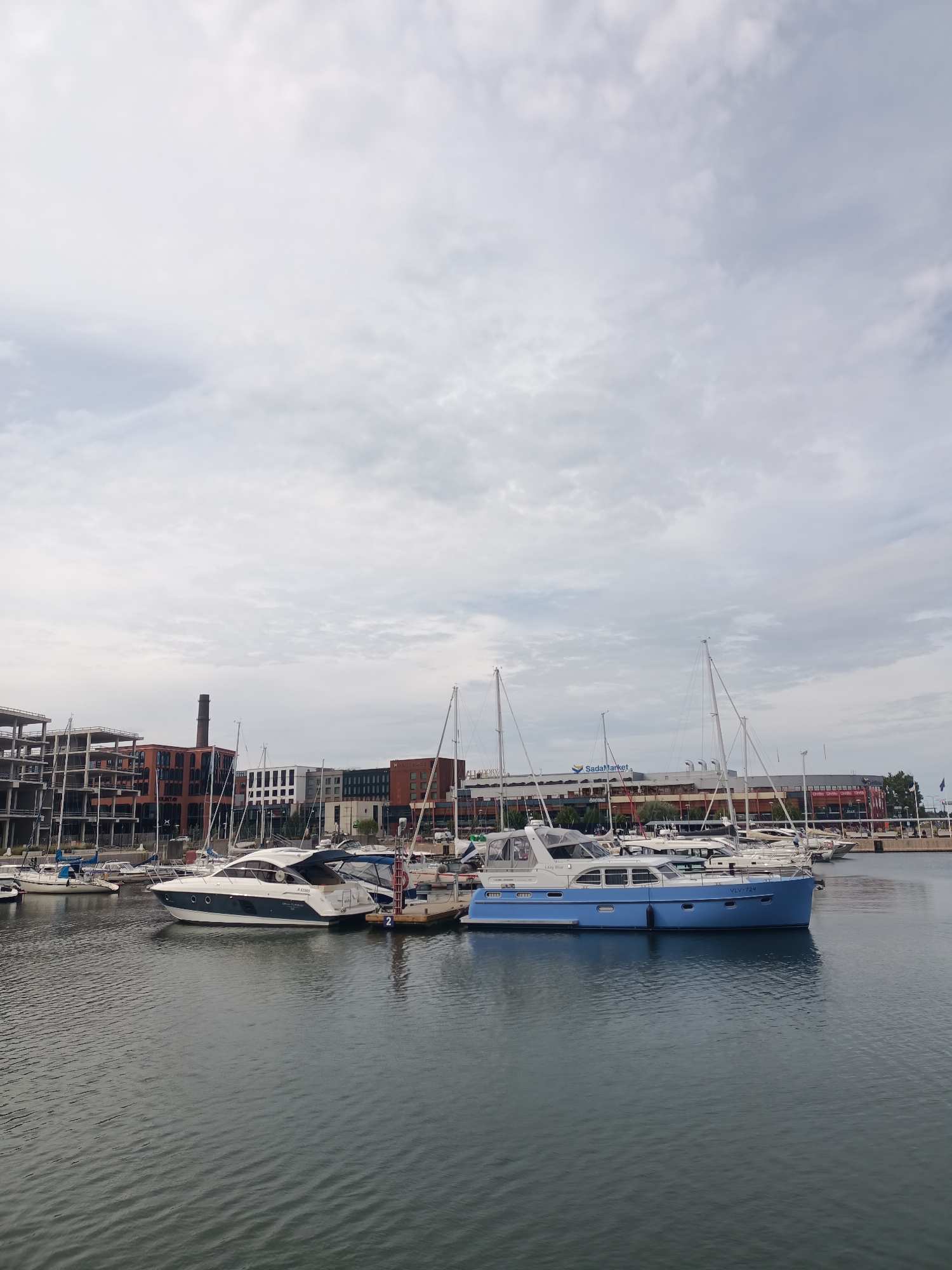 The image depicts a marina in Tallinn, Estonia, with several boats docked. In the background, there are modern buildings, including what appears to be a shopping center with the "SadaMarket" sign visible, and older brick structures with smokestacks, suggesting a redeveloped industrial area. The sky is overcast with clouds. The foreground is dominated by the water of the marina, with the hulls of several yachts and motorboats visible. One prominent motorboat in the center of the frame is light blue and white, with the registration "VLV-724" visible. Another white and dark blue motorboat is docked next to it. A few sailboats with tall masts are also present in the marina. The overall scene is one of a calm day at the harbor, with urban infrastructure and maritime activity.