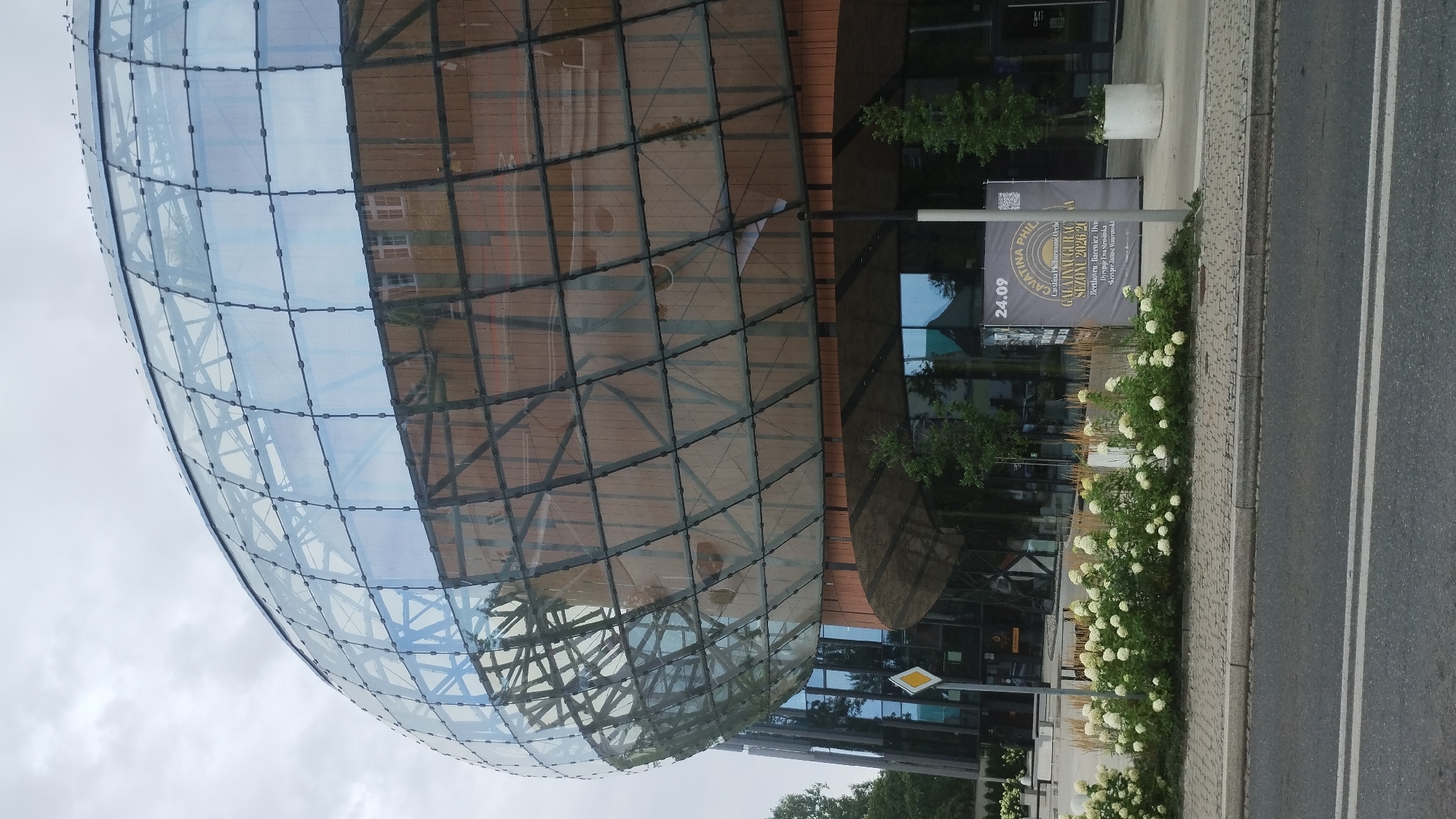 The image shows a modern building with a large, curved glass facade supported by a dark metal structure. The building is likely located in Bielsko-Biala, Poland, as indicated by the sign visible on the right. The sign, which appears to be a banner or poster, displays the text "24.09 CAVATINA PHIL GALA INAUGURAC SEZONU 2026/20" and mentions "Beethoven," suggesting it is an announcement for a philharmonic concert or season opening.
In front of the building, a sidewalk runs along a street. A paved sidewalk is lined with a decorative garden bed filled with white flowering bushes and tall ornamental grasses. A gray street with white lane markings is visible in the foreground. A traffic sign with a yellow diamond shape is positioned on the left side of the sidewalk. The sky is overcast and gray. No people are visible in the image. The architectural style of the building is contemporary and futuristic, characterized by its spherical shape and extensive use of glass.