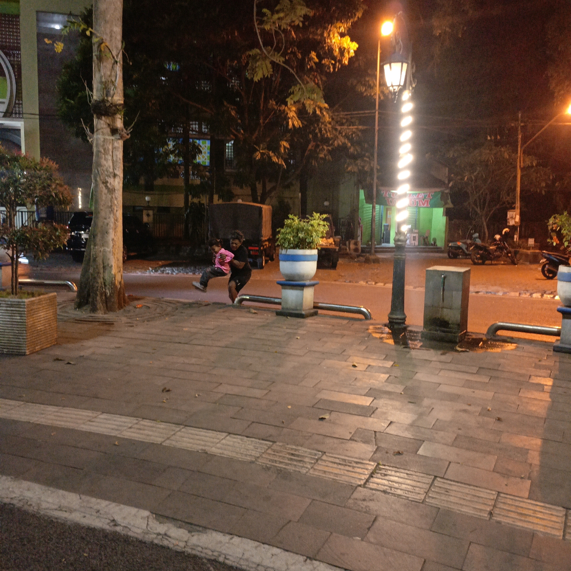 The image depicts a night scene on a paved sidewalk in Salatiga, Indonesia. Two individuals are engaged in playful activity. One person, wearing a pink shirt and dark pants, is being lifted and swung by another person in a black shirt and shorts. The background shows a street with parked motorcycles and a building with green signage. A lamppost with decorative lights illuminates the scene, casting shadows on the wet pavement. A large tree trunk is visible on the left side of the frame.