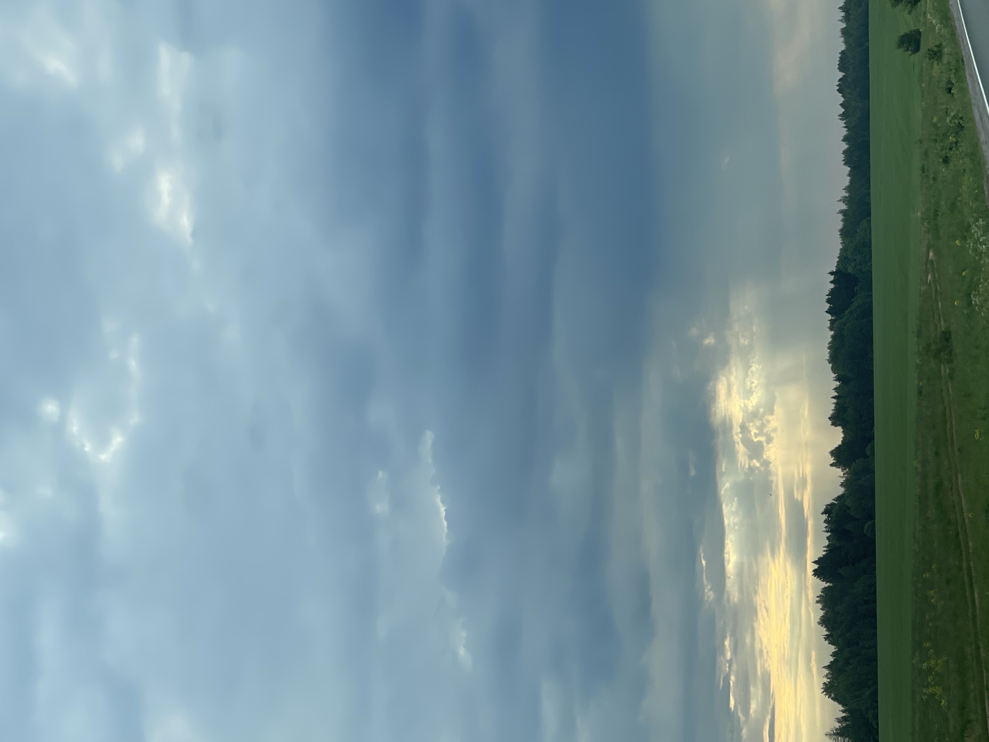 This media file captures a serene landscape dominated by a vast, cloudy sky. The image depicts a wide expanse of sky, filled with a mix of light blue and various shades of grey clouds, indicating a dynamic weather pattern. Towards the right side of the frame, the sun peeks through a break in the clouds, casting a warm, golden light that illuminates the edges of the clouds and creates bright, ethereal streaks.
Below the expansive sky, a distinct horizon line separates the heavens from the earth. A dark, dense line of trees forms a forest, stretching horizontally across the landscape, silhouetted against the sky. Adjacent to the forest, a vibrant green field or grassland extends into the foreground. On the far right edge, a narrow, light-colored strip is visible, suggesting a dirt track or a distant road running alongside the field.
The time of day appears to be late afternoon or early evening, possibly around sunset, given the warm, golden hue of the sunlight breaking through the clouds. The setting is a natural, rural environment, devoid of any specific location cues like landmarks or buildings. No people or human-made objects are visibly present, nor is there any discernible activity taking place. The overall atmosphere conveyed is one of peacefulness and natural beauty. There is no visible text in the image.