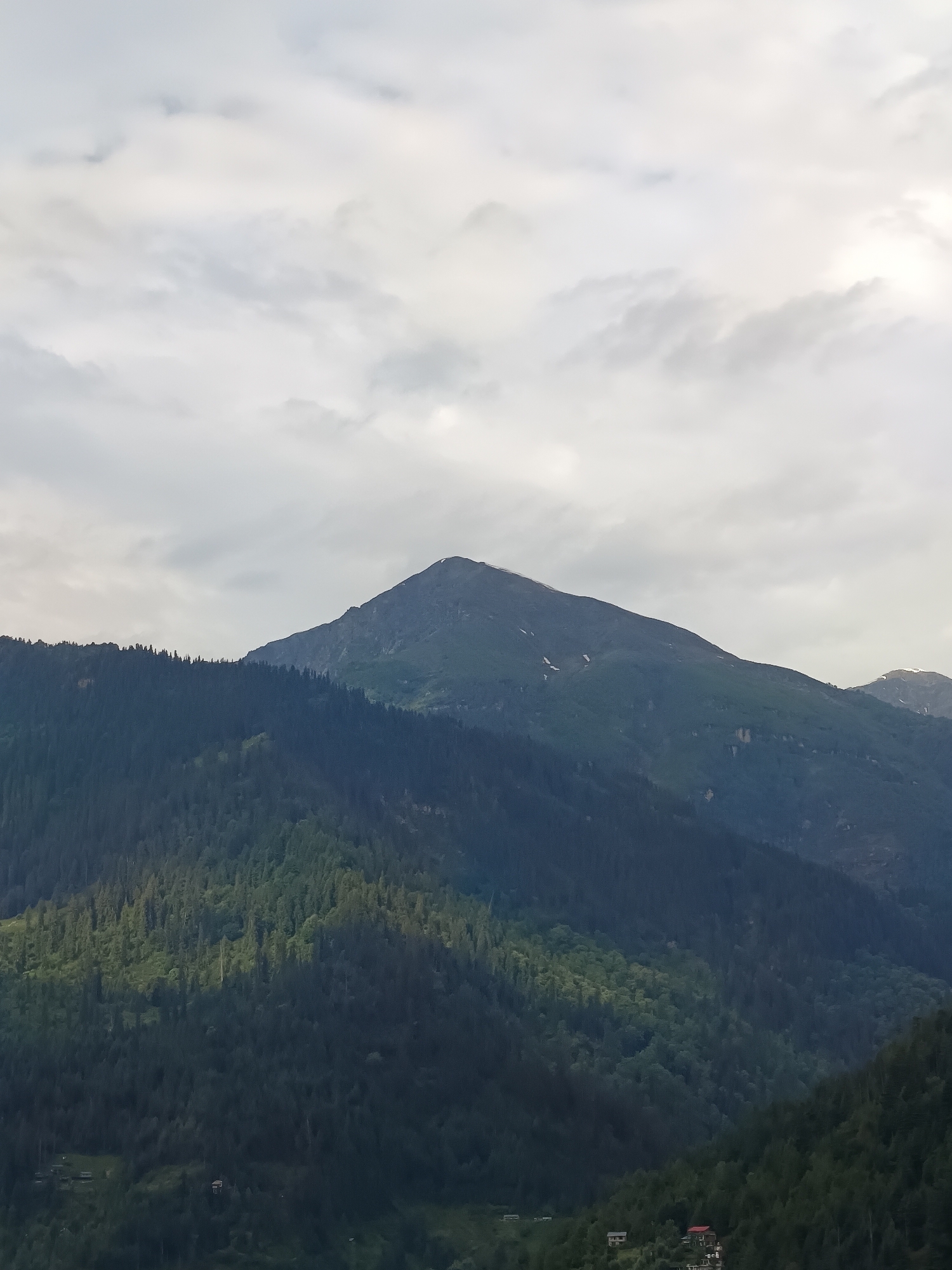 The image shows a wide mountain landscape with a prominent peak in the center, against a cloudy sky. The lower slopes of the mountains are covered in dense evergreen forest. Towards the bottom of the frame, there are some small structures and buildings scattered amongst the trees, indicative of a rural settlement. The overall scene is natural and serene.