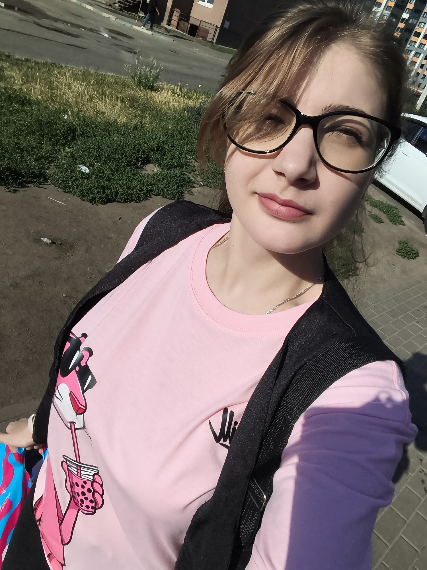 A young woman is taking a selfie outdoors, standing on a sidewalk. She has blonde hair and is wearing black-rimmed glasses, a pink t-shirt with a cartoon panther graphic, and a black vest or jacket over her shoulders. The background shows a street, some green grass and bushes, and a tall apartment building. The sun is shining, casting shadows.
