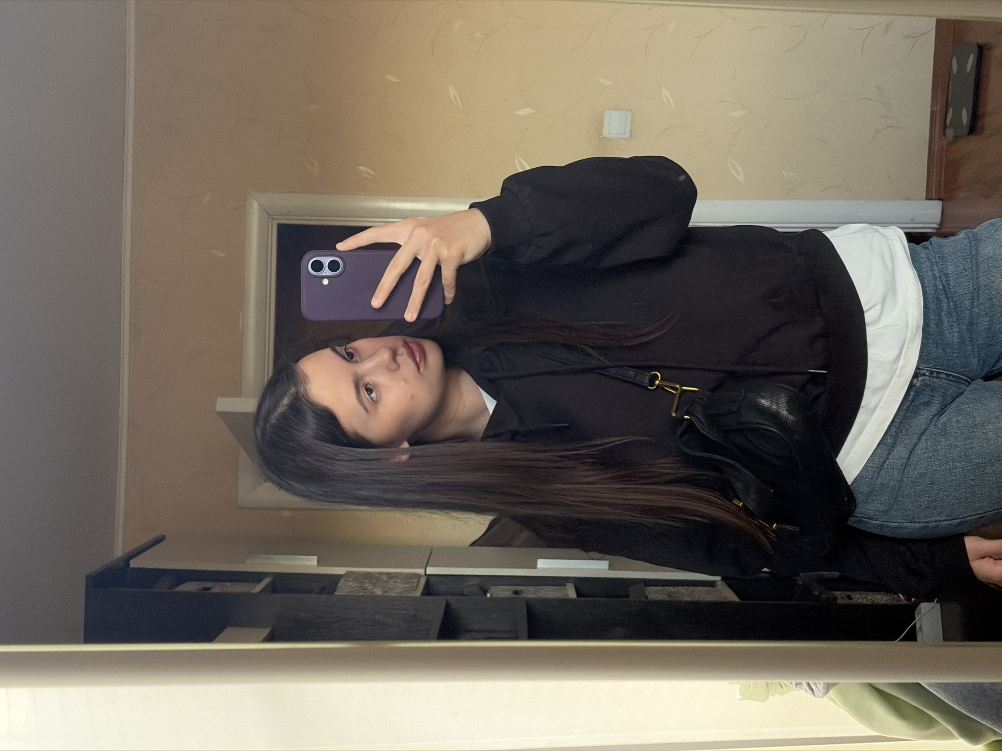 A young woman is captured taking a mirror selfie indoors. She has long, straight dark brown hair and fair skin, with a neutral expression, looking directly into the camera's reflection. She is wearing a black hooded sweatshirt over a white undershirt and blue jeans. A black shoulder bag with gold-toned hardware is visible draped across her.
She holds a purple smartphone, which appears to be an iPhone model with dual rear cameras, up to take the photo. The setting is an indoor space, likely a room or hallway in a residence. The background wall features light beige wallpaper with a subtle, delicate leaf or vine pattern. A white electrical outlet is visible on the wall, and a light-colored doorframe is reflected in the mirror. Below the woman's reflection, parts of a dark, multi-compartment organizer are visible on what appears to be a shelf or cabinet. Further down, reflections of light green and grey fabric, possibly clothes, can be seen. The ambient lighting suggests it is daytime. The location context provided indicates this scene is in Chaikovsky, Russia. No visible text is present in the image.