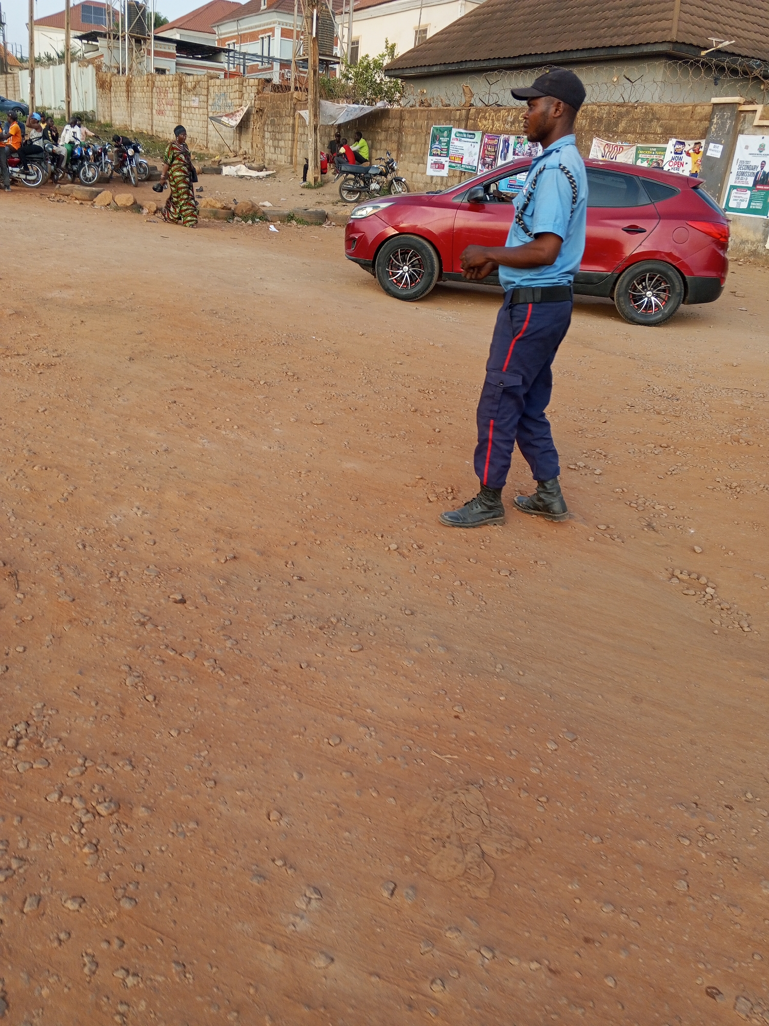 The image depicts a man in a security uniform standing on a dirt road in Abuja, Nigeria. The man is wearing a blue short-sleeved shirt, dark blue trousers with a red stripe, black boots, and a black baseball cap. He has a rope-like object across his chest. Behind him is a red SUV parked on the side of the road. Further back, there are motorcycles, pedestrians, and buildings under a partially cloudy sky. Advertisements are visible on the wall behind the vehicle. The ground is reddish-brown dirt and scattered with small stones.