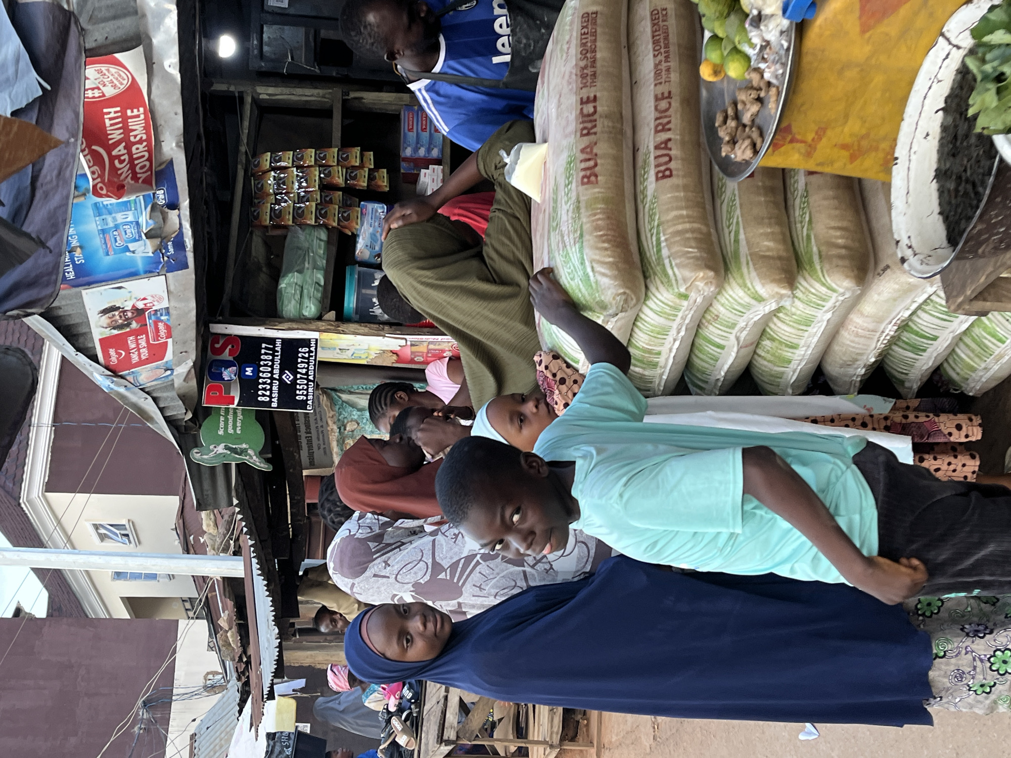 The image depicts a bustling marketplace scene in Jos, Nigeria. In the foreground, a young boy wearing a light blue t-shirt and dark pants faces the camera, his expression playful. To his left stands a young girl in a dark blue hijab and patterned skirt. Behind them, several other people are visible, including a woman in a brown hijab, another in a white and brown patterned headscarf, and a person in a green garment.
The marketplace appears to be a street stall with a corrugated metal roof. Signs are displayed, including a red Colgate advertisement with the text "YANGA WITH YOUR SMILE" and another smaller sign with "Colgate Oral B." Further signs indicate prices or business names, with partial text visible such as "POS" and numbers. Stacks of large rice bags, labeled "BUA RICE 100% SORTEXED THAI PARBOILED RICE," are prominently displayed. To the right, a man in a blue and white striped shirt and a black apron is partially visible, suggesting he is the vendor. A metal tray on a yellow surface holds ginger and limes. Other items, possibly spices or herbs, are in a white basin in the lower right corner. The overall environment suggests an active commercial area with goods for sale.