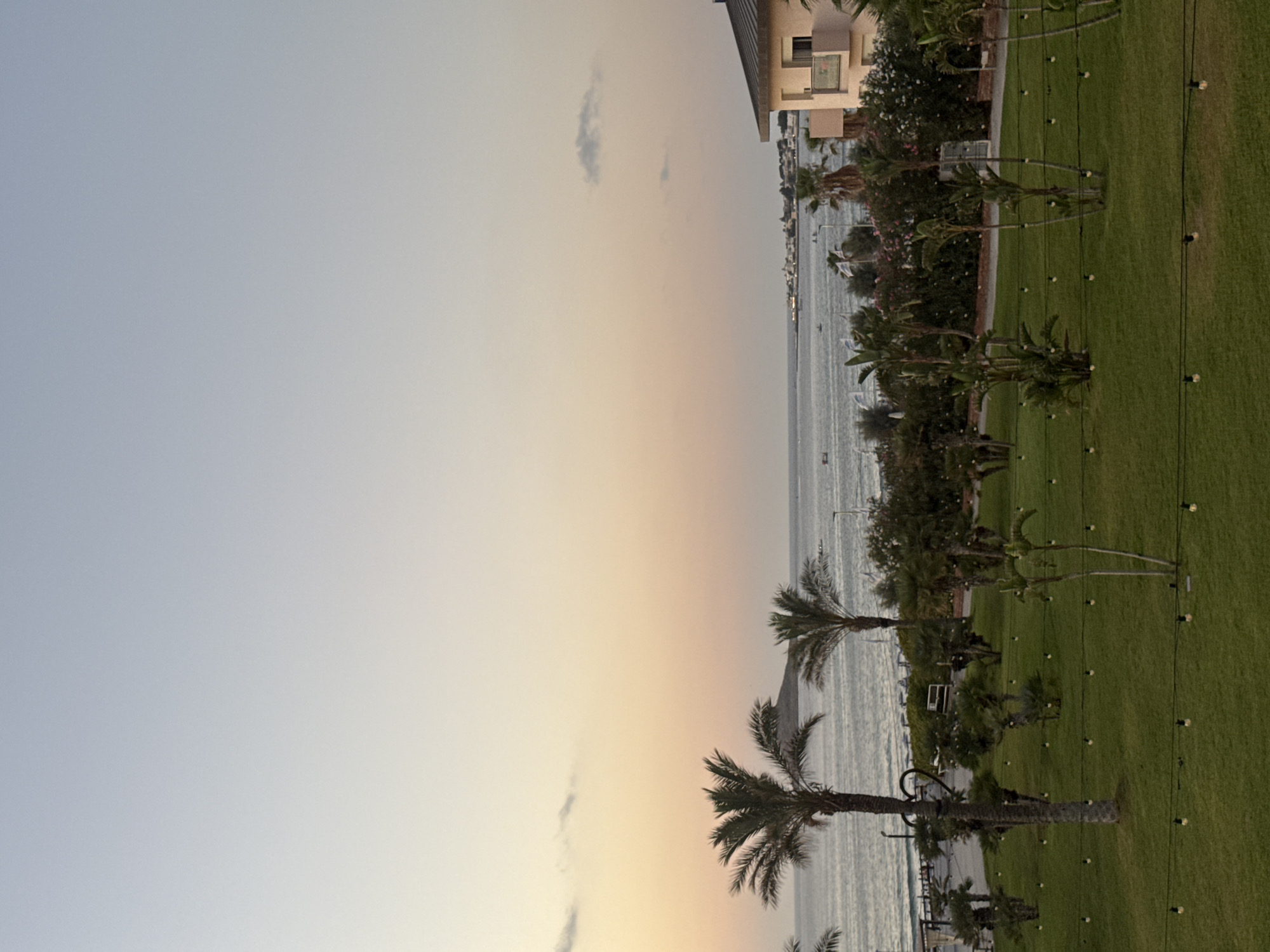 The image captures a serene sunset scene in Isola delle Femmine, Italy, overlooking the sea. The sky transitions from a soft blue at the top to warm hues of peach and yellow near the horizon, with a few wispy clouds scattered across. In the foreground, a lush green lawn is adorned with strings of small, illuminated light bulbs, creating a festive ambiance. Several palm trees, some tall and slender, others shorter and bushier, are interspersed across the lawn and a paved pathway.
Beyond the lawn, a body of water, likely the Mediterranean Sea, stretches to the horizon. The water is calm, with gentle waves visible. A distant island or landmass with a hill can be seen to the left of the frame. Along the coastline, a row of buildings, including a pink multi-story structure with balconies on the right, marks the edge of the town. Boats are visible on the water, some small and appearing to be moored, while others seem to be in motion. The overall atmosphere is tranquil and picturesque, characteristic of a coastal Italian town at dusk.