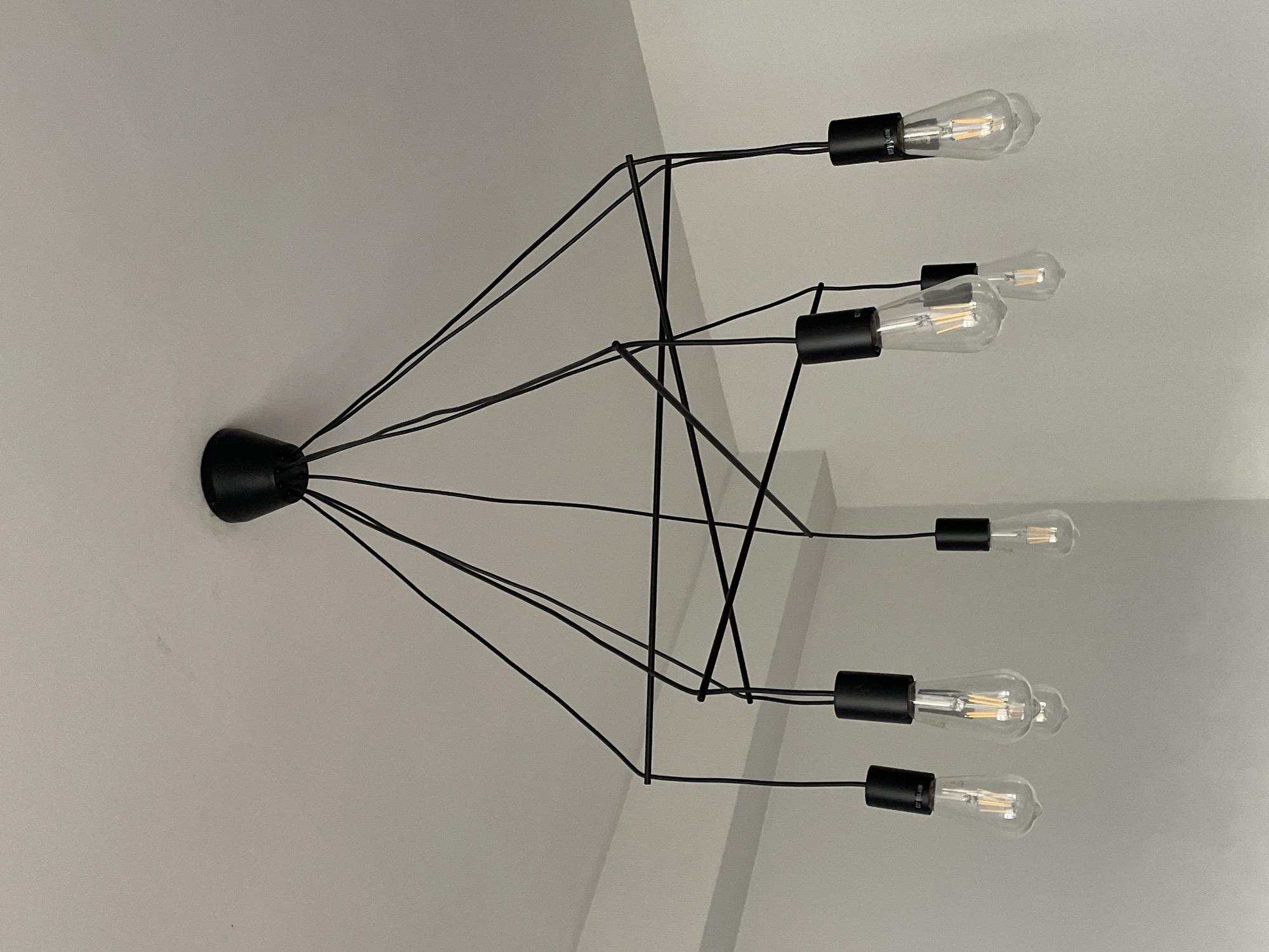 A black, contemporary chandelier with exposed Edison bulbs is mounted on a light gray ceiling. The fixture consists of a central black base from which multiple black wires extend to hold individual bulbs. The bulbs are elongated and clear, revealing their filaments. The background is a neutral, undecorated interior. The location is not discernible from the image.