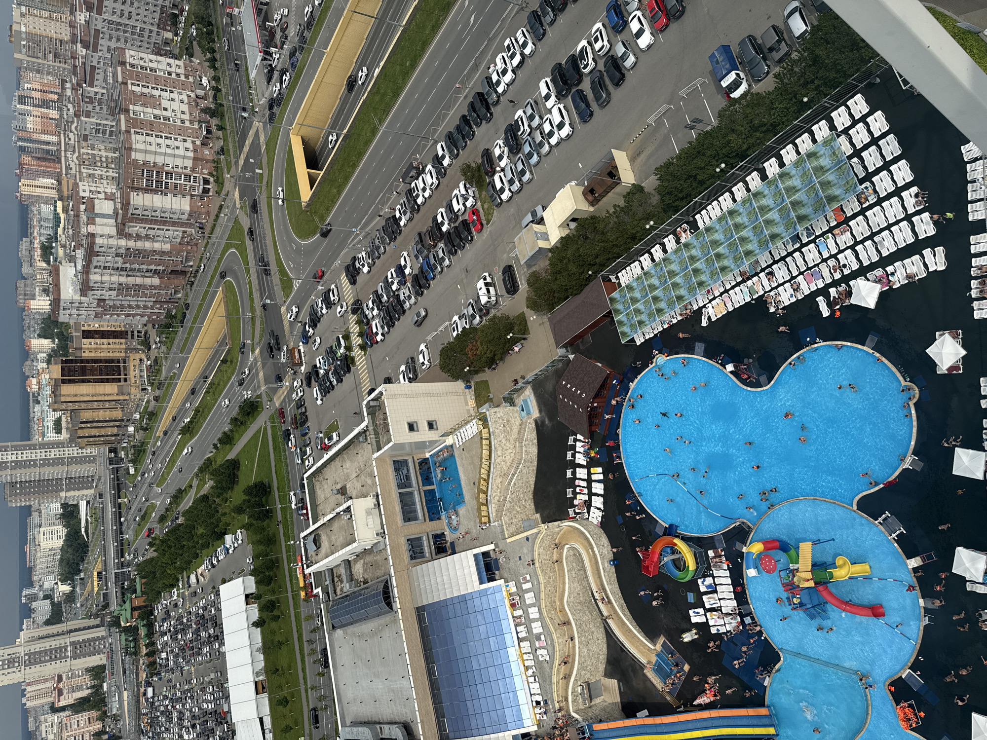 The image captures an aerial view of a water park, likely located in Kazan, Russia, as indicated by the architectural style of the surrounding residential buildings and the presence of a large, multi-lane road infrastructure typical of urban areas. The water park itself features several large, irregularly shaped swimming pools filled with bright blue water. One prominent pool has a central children's play area with colorful slides, climbing structures, and spray features. Another larger pool is populated with numerous people swimming and relaxing. Surrounding the pools are extensive sunbathing areas with rows of white lounge chairs, some covered by canopies. Adjacent to the water park, there is a large parking lot filled with many cars. Further beyond the water park and parking lot, a dense urban landscape unfolds, characterized by tall, modern residential buildings, a multi-lane highway with circulating traffic, and some commercial structures. The sky is overcast, suggesting a cloudy day.
