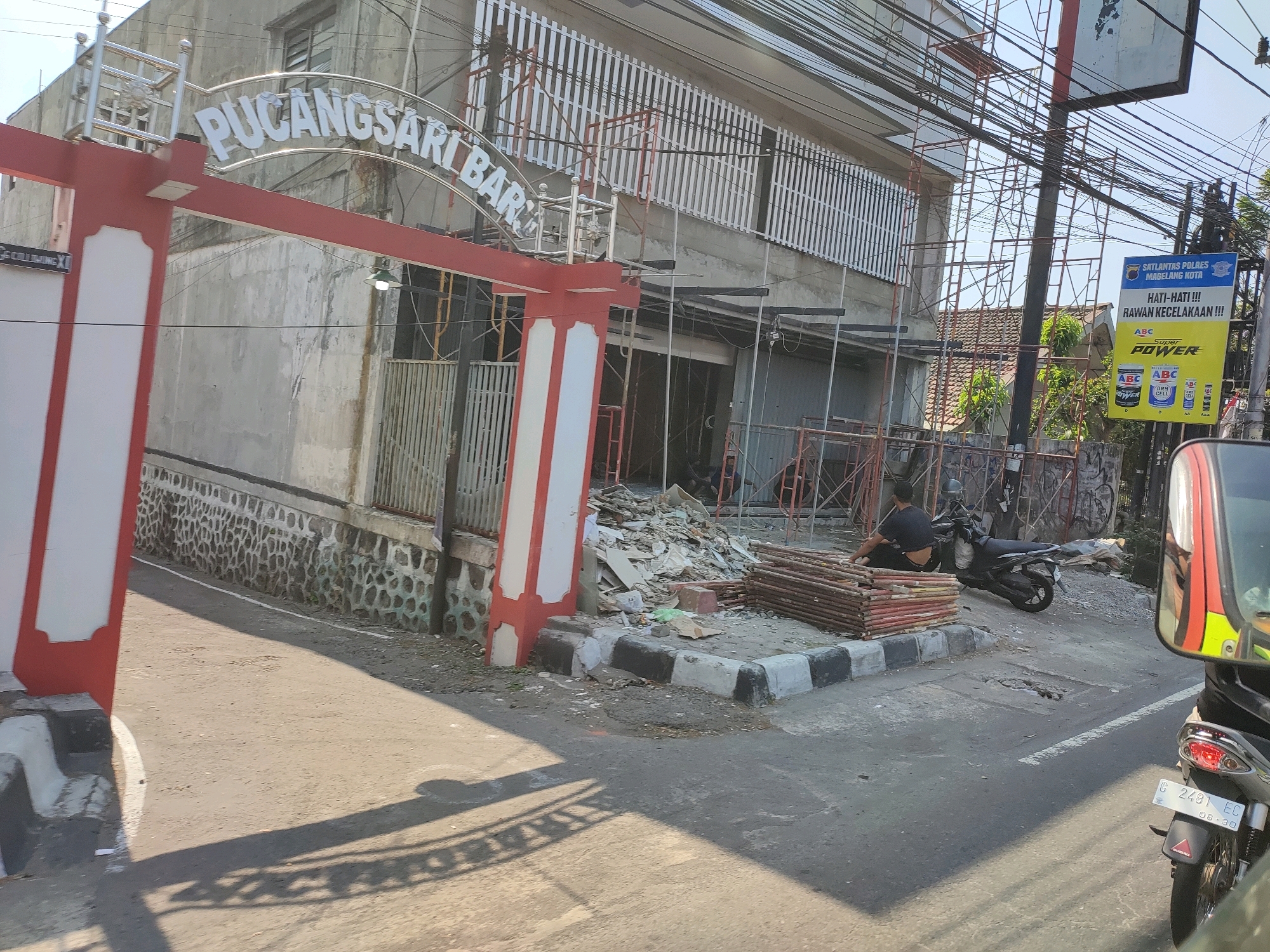 The image shows a street scene in Magelang, Indonesia, with a building under construction or renovation. A red and white archway with the words "PUCANGSARI BAR" is prominent in the foreground, partially obscuring the building. Scaffolding surrounds the building, and construction materials, including piles of wood, are scattered on the ground. A person is sitting on a stack of wood near a motorcycle. A blue and yellow billboard with text in Indonesian, including "HATI-HATI !!! RAWAN KECELAKAAN !!!" (Be Careful!!! Accident Prone!!!), is visible on a pole. The street surface appears to be asphalt, with a curb and sidewalk. Shadows from overhead wires and the archway are cast on the ground. A motorcycle is parked on the right side of the frame, with its license plate visible, reading "G 2481 EC". The sky is clear and blue.