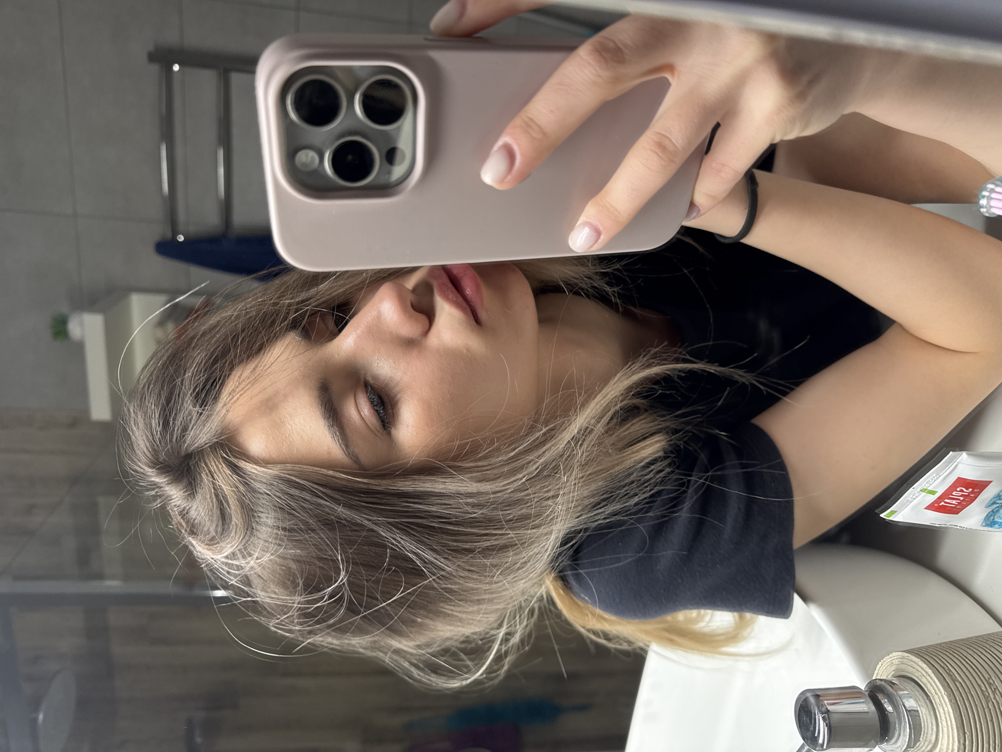 A young woman is taking a selfie in a bathroom mirror. She is holding a pink smartphone in her right hand, and her face is partially obscured by the phone. She has light brown hair and is wearing a black shirt. The bathroom contains a towel rack, sink, toilet paper, and a package of wipes. The location is implied to be Chaikovsky, Russia.