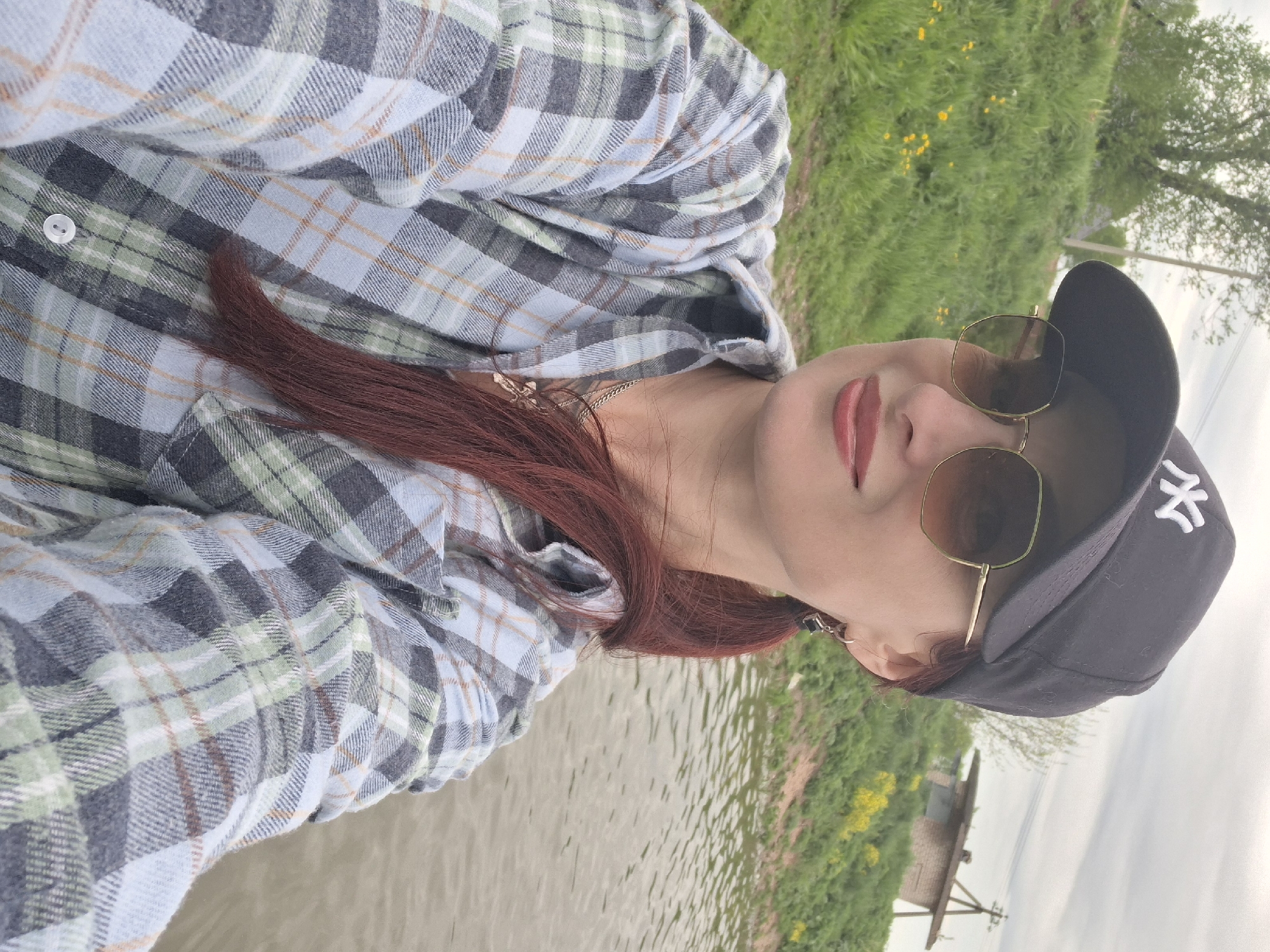 A woman is photographed outdoors in what appears to be the Забалоцкі сельскі Савет area in Belarus. She's wearing a plaid shirt, sunglasses, and a dark baseball cap with a white logo. Her hair is reddish-brown. The background includes greenery, possibly grass and trees, along with a body of water. A simple structure is also visible in the distance.