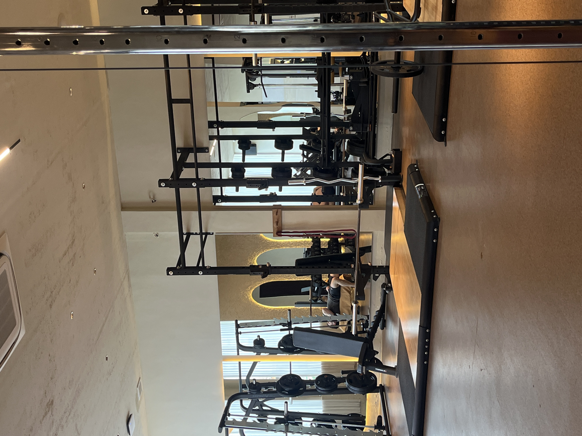The image shows the interior of a gym. Several weightlifting racks and benches are visible, indicating a space designed for strength training. Mirrors are present, reflecting the equipment and creating an illusion of depth. Someone appears to be working out on one of the benches, seen through the reflection. The environment is a gym, likely in North Kuta, Indonesia.