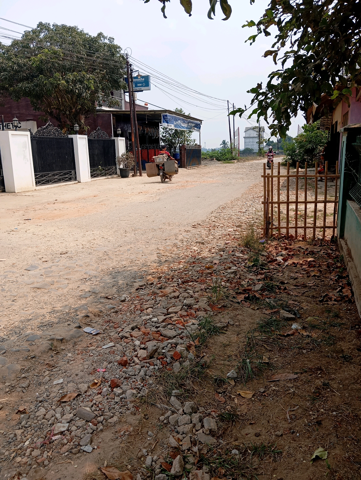 The image depicts a street scene in Batang Hari Ogan, Indonesia, likely a rural or semi-rural area. The road is unpaved and appears to be made of gravel and dirt, with some visible cobblestones in the foreground. The right side of the road is lined with a low bamboo fence and some foliage, including dry leaves scattered on the ground. To the left, a white fence with black wrought iron accents encloses a property with a building behind it.
A person on a motorcycle is visible in the middle distance, traveling away from the camera. The motorcycle appears to be carrying a load secured at the back. Further down the road, a large tree with dense foliage stands to the left, near a building with a sign that reads "Annoche." Another building with a blue sign is also visible in the distance. Utility poles with numerous wires are present along the road. In the far distance, a water tower can be seen. The sky is overcast. The overall impression is of a somewhat dusty and undeveloped road.