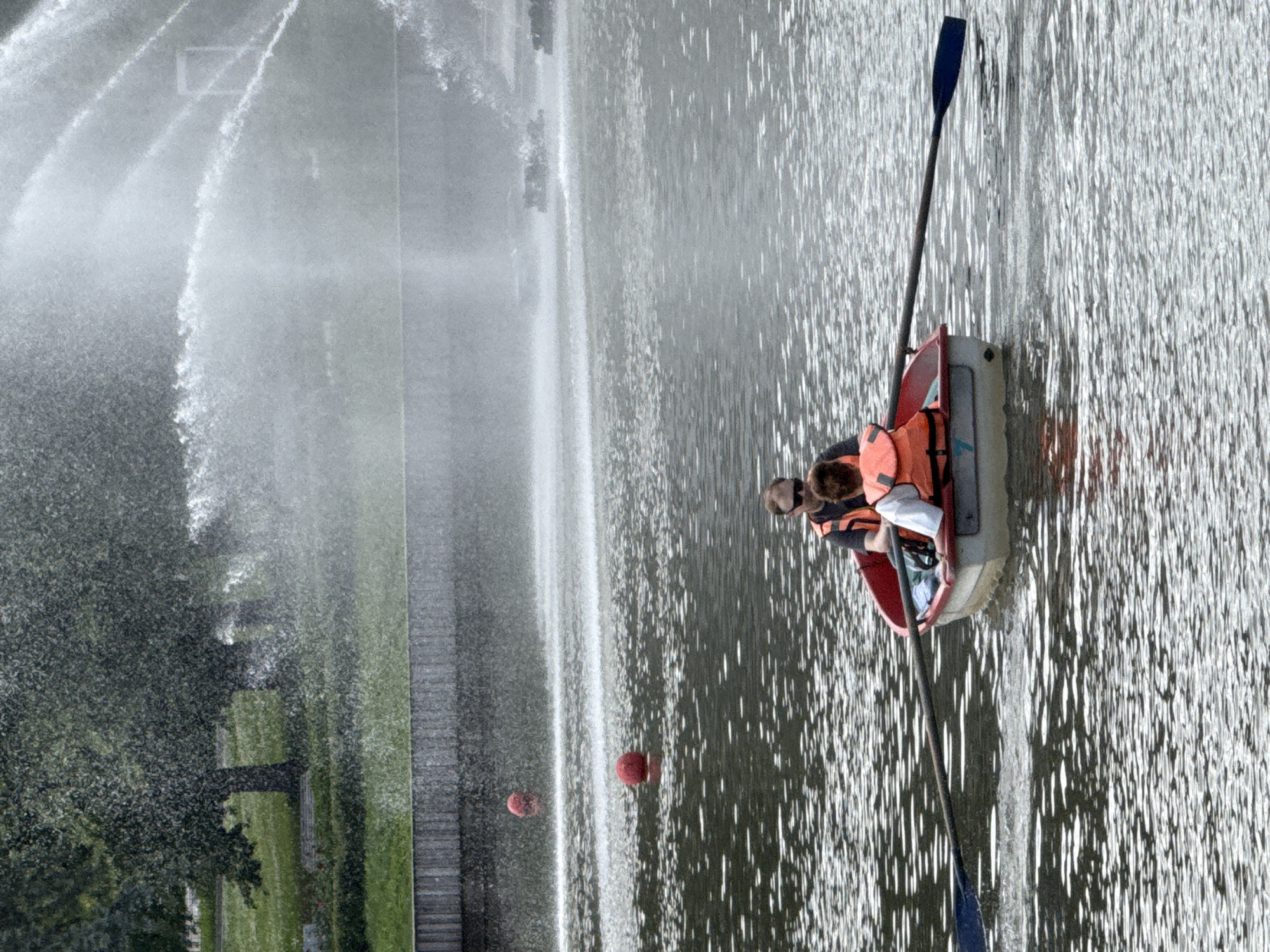 Two individuals are in a rowboat on a body of water, with a fountain spraying water in the background. The person in the front is steering with an oar, while the person in the back is looking forward. Both individuals are wearing life jackets. The water is choppy, and there are two red buoys in the water. The scene appears to be in a park or recreational area, possibly located in Moscow, Russia. The fountain is large and is spraying water high into the air, creating a misty effect. Trees and grass are visible on the bank behind the fountain. The overall impression is one of a leisurely day on the water.