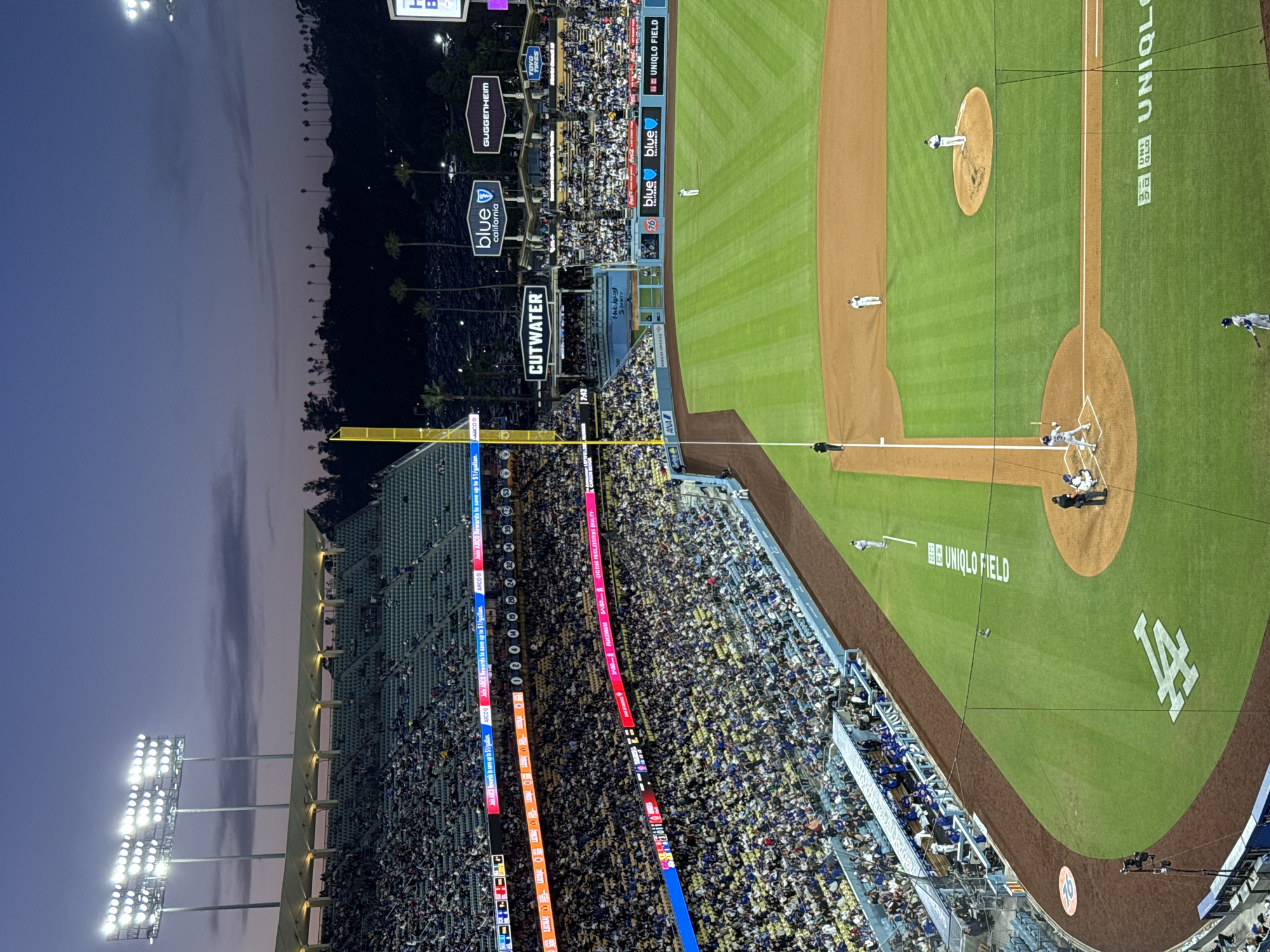 This is an eye-level, rotated photograph of Dodger Stadium located in Los Angeles, California, United States of America. The image is set during twilight.
In the foreground, there are baseball players on the field near home base. A batter stands ready while the catcher crouches behind him. Two other players stand nearby. The field displays "UNIQLO FIELD" and the "LA" logo.
The middle ground is filled with a vast crowd of spectators in the stadium seating, divided by levels of seating, aisles and walkways. Signage includes advertisements such as "ARCO", "CUTWATER", "blue california", "GUGGENHEIM", "TOYO TIRES", and "UNIQLO FIELD". The seating extends upwards and into the background.
The background contains stadium lights on the left, palm trees, and a darkening sky.
