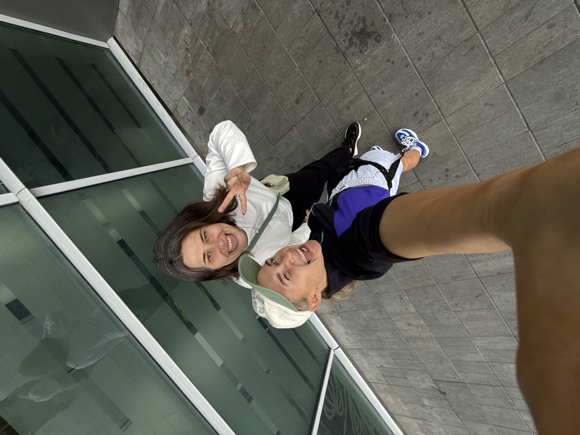 Two individuals are posing for a selfie outdoors. The person in the foreground, wearing a black jacket, purple and blue accents, light grey shorts, and a light-colored baseball cap, is sticking their tongue out at the camera. The second person, standing behind and slightly to the right, is wearing a white long-sleeved shirt, black pants, and is making a peace sign with their right hand. They are both smiling. The background consists of a modern building with large, dark green windows and a light-colored frame, and a paved walkway made of grey rectangular tiles. The overall scene suggests a casual, friendly interaction in an urban or institutional setting. There is no visual information in the image to suggest the location is Bol'shie Kabany, Russia.